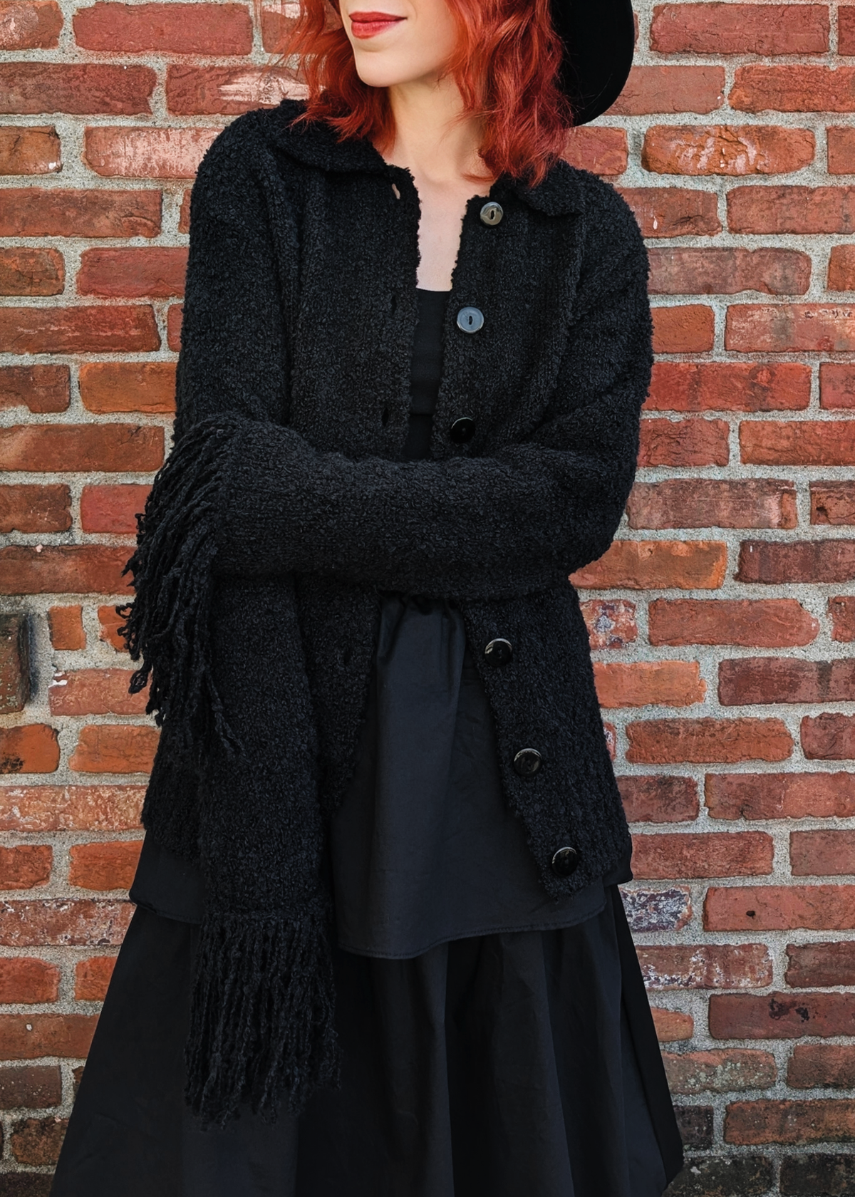 Person wearing a black cardigan with fringe cuffs against a brick wall
