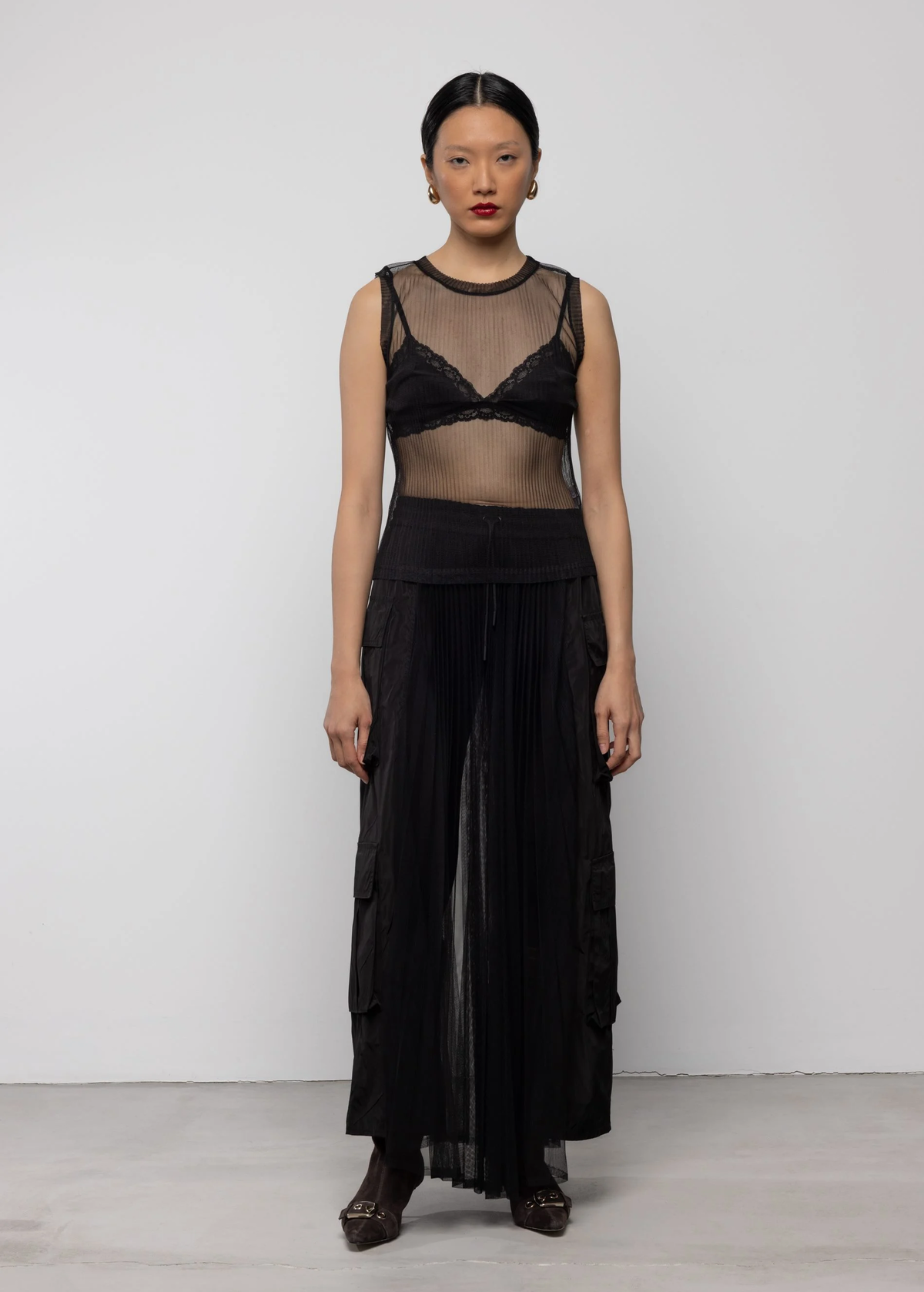 Woman wearing a black sheer top and long black sheer pleated cargo skirt against a white background