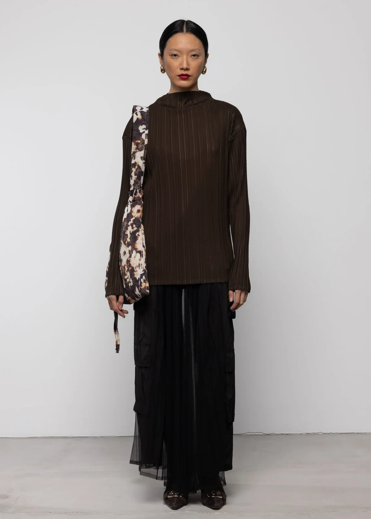 Woman wearing a brown ribbed top and black sheer mesh cargo skirt with a floral purse over her shoulder against a white background