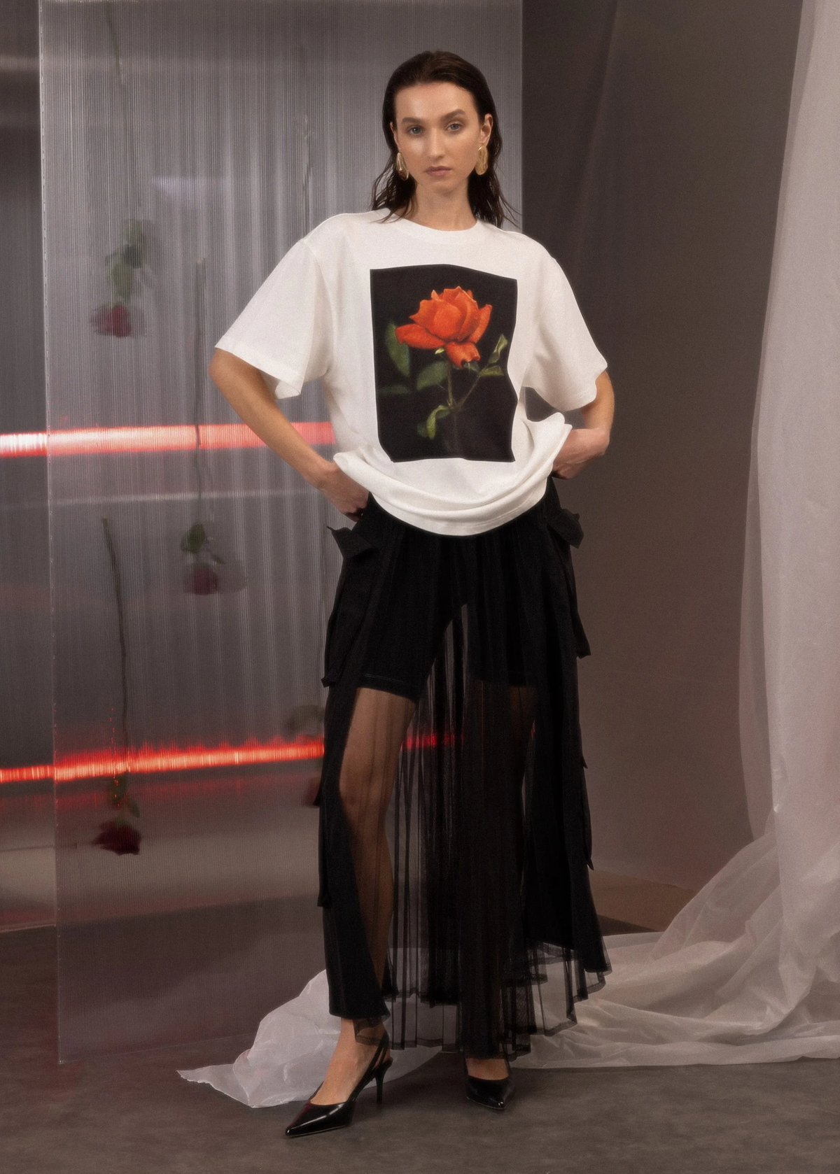 Woman wearing a white t-shirt with a rose graphic and a black skirt in a minimalistic setting.