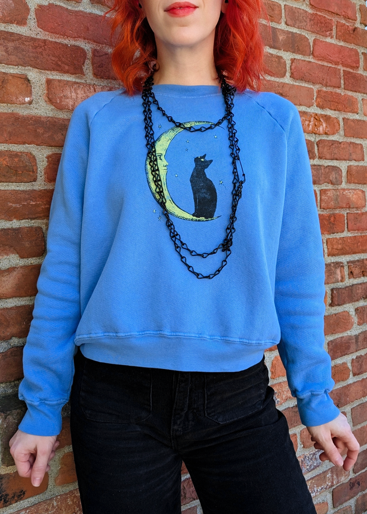 A person with bright red hair wearing a light blue crewneck sweatshirt with a black cat on a crescent moon graphic, styled with layered black chain necklaces and black jeans, standing against a red brick wall.