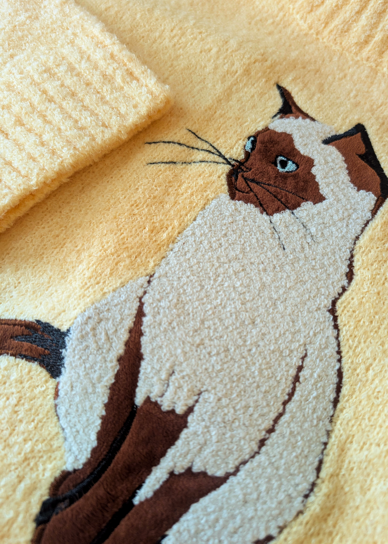 Close-up of a butter yellow-colored knit fabric featuring an embroidered cat motif in brown and cream with blue eyes