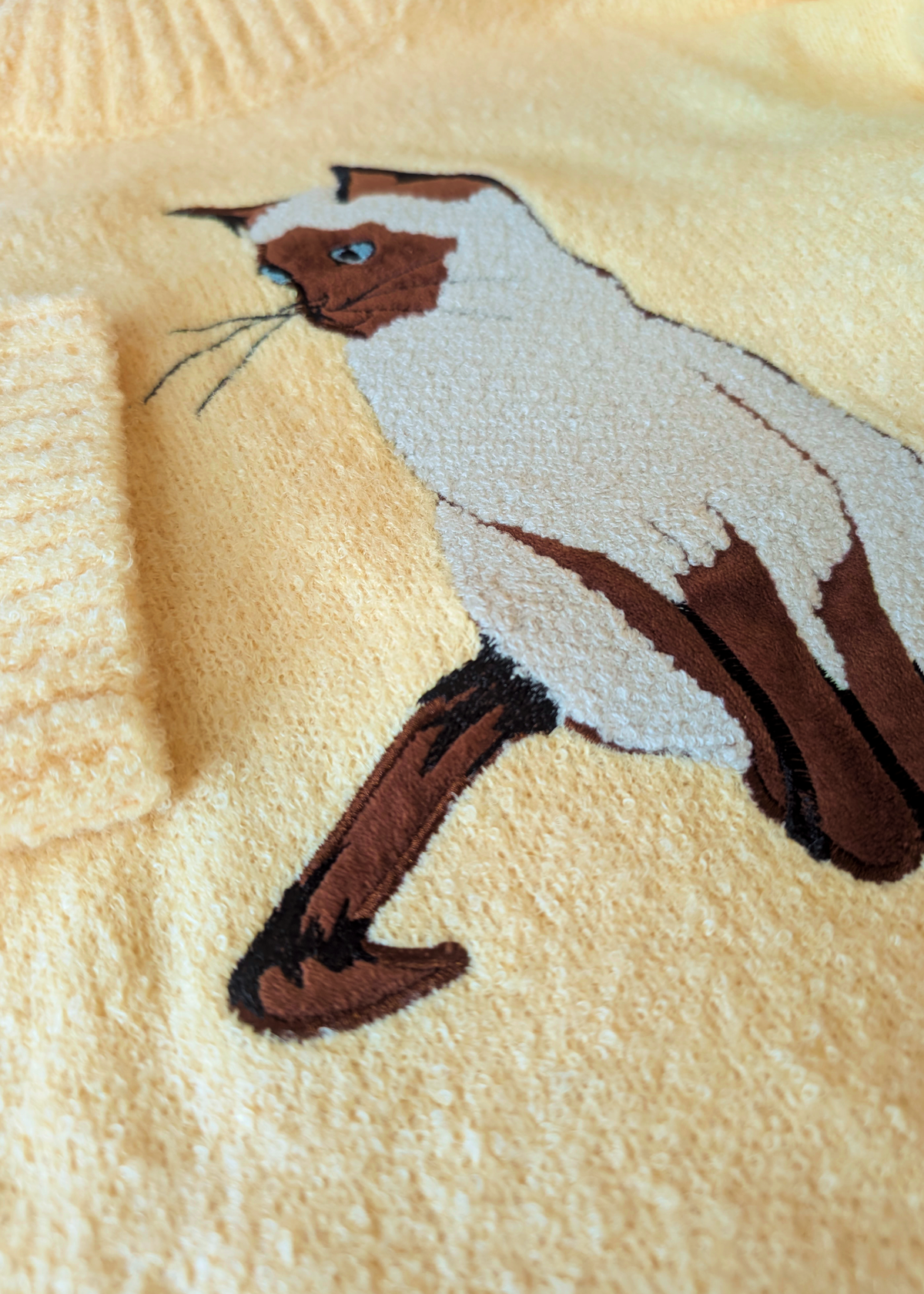 Close-up of a yellow-colored knit sweater featuring an embroidered Siamese cat design on the front.