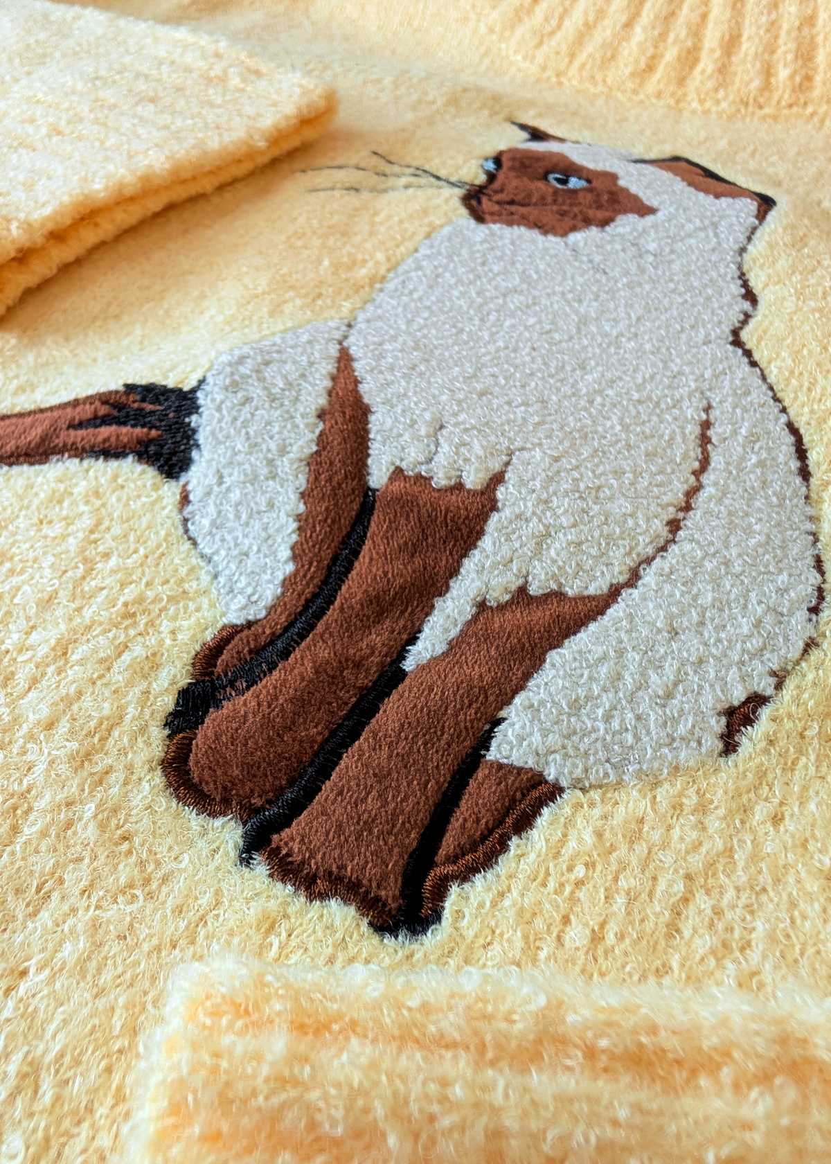 A close-up of a textured yellow sweater featuring a raised, tufted embroidered cat design in brown, cream, and black.
