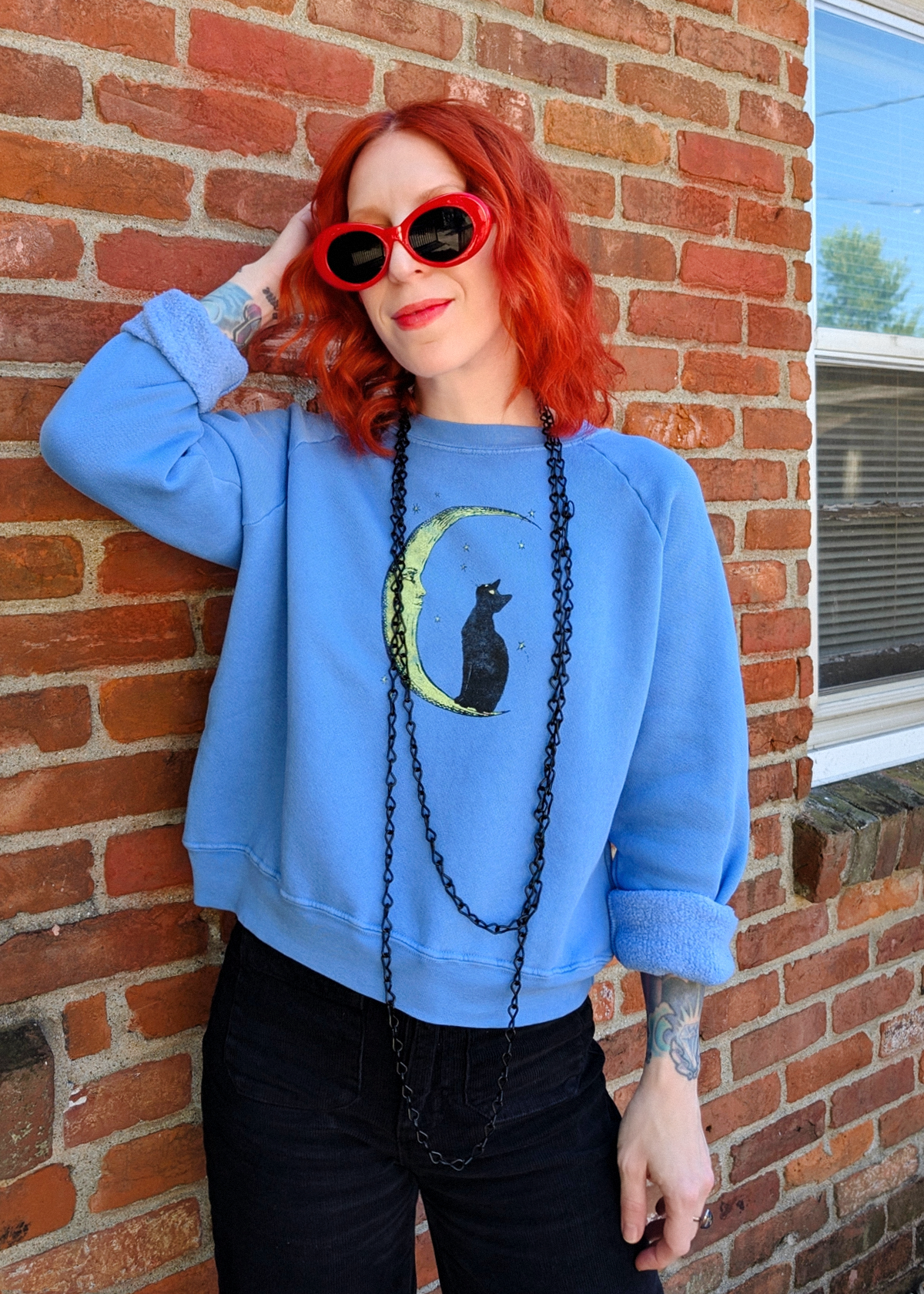 A woman with bright red hair and red sunglasses leans against a red brick wall, wearing a light blue crewneck sweatshirt with a black cat on a crescent moon graphic, black jeans, and a long black chain necklace.