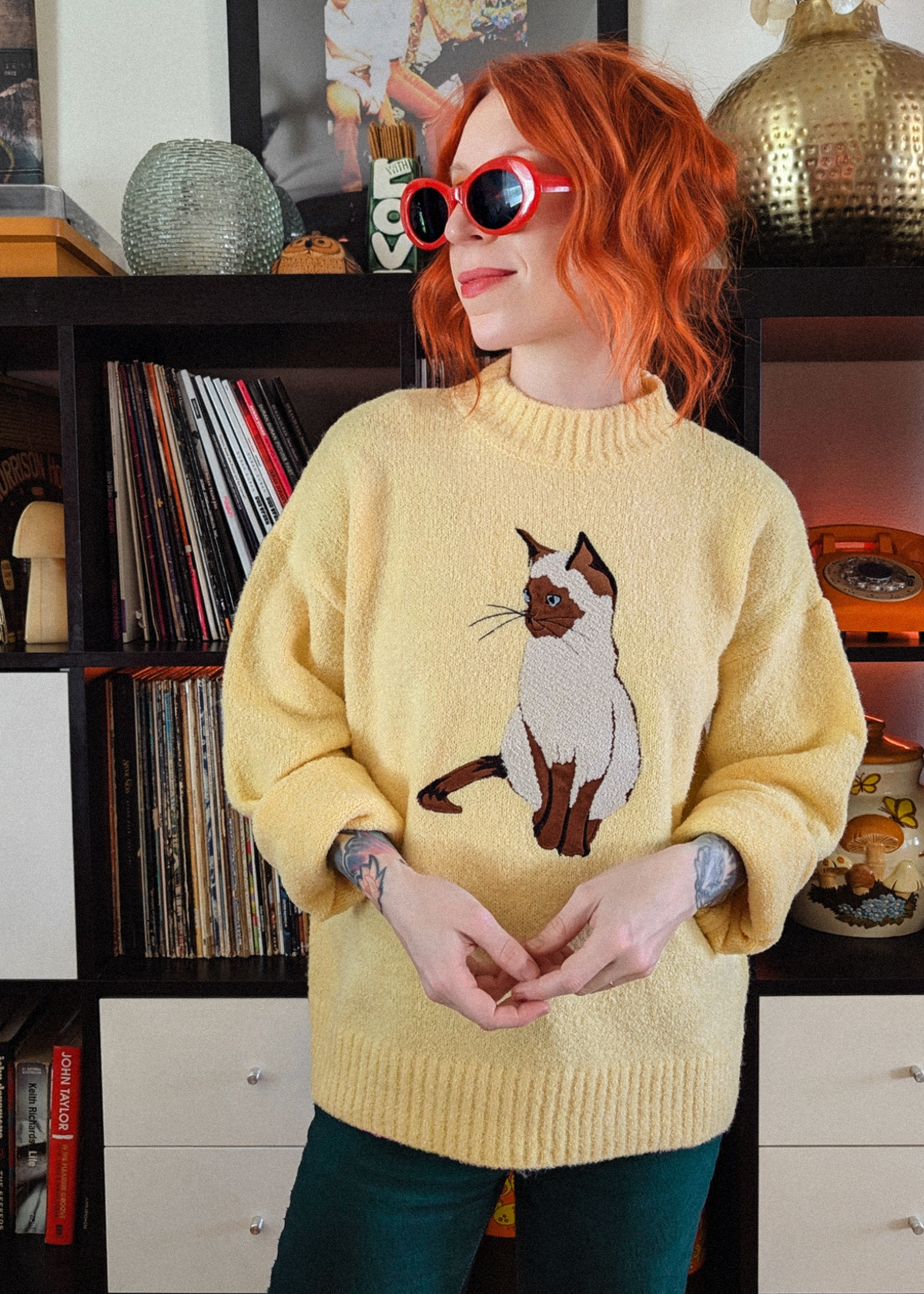 A person with red hair wearing a fuzzy pale yellow knit sweater with a large cat graphic on the front, paired with dark green pants and red sunglasses, standing in front of a bookshelf.