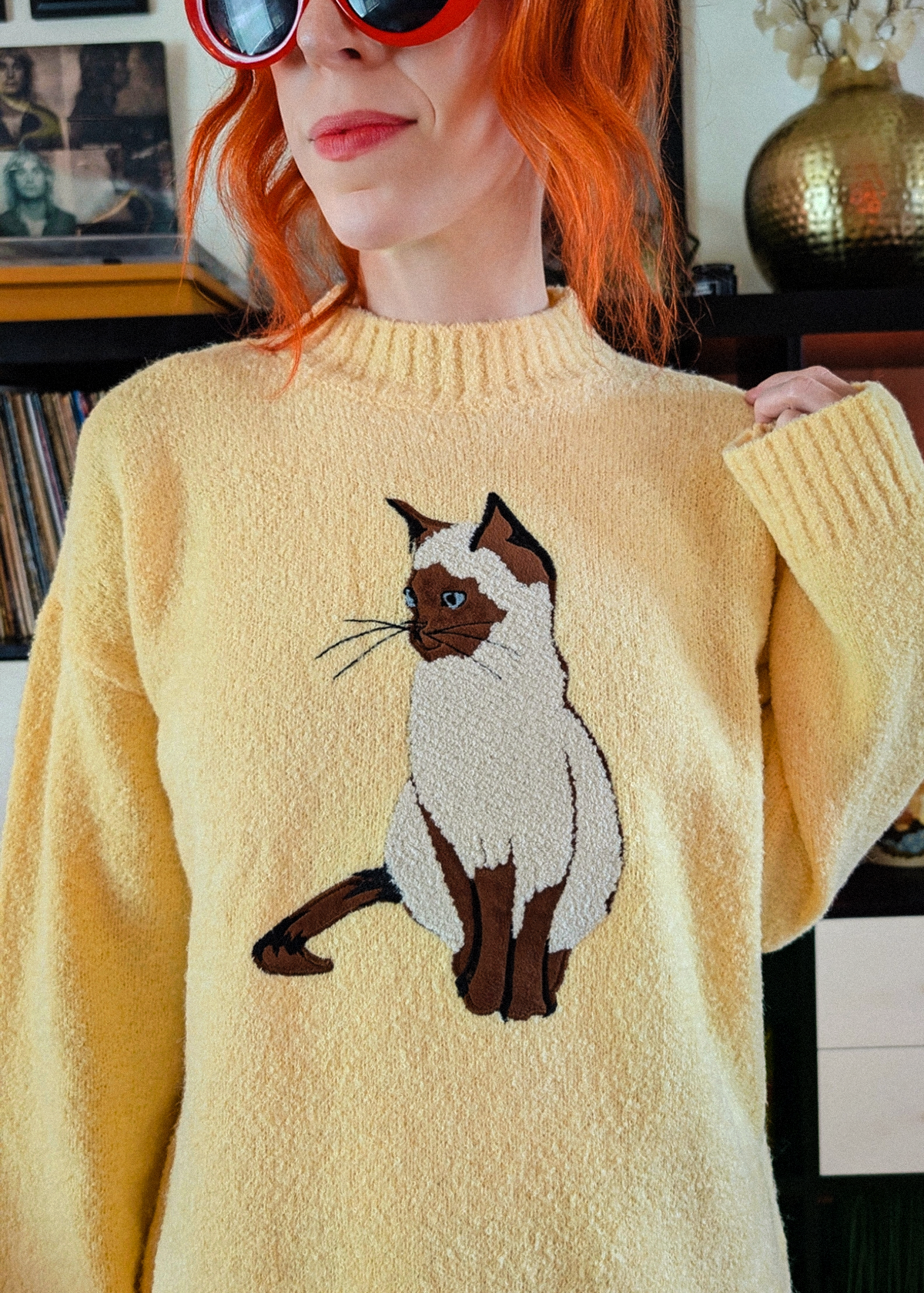 A person with bright red hair wearing a fuzzy pale yellow sweater with a large embroidered cat motif on the front, photographed indoors with a blurred background.