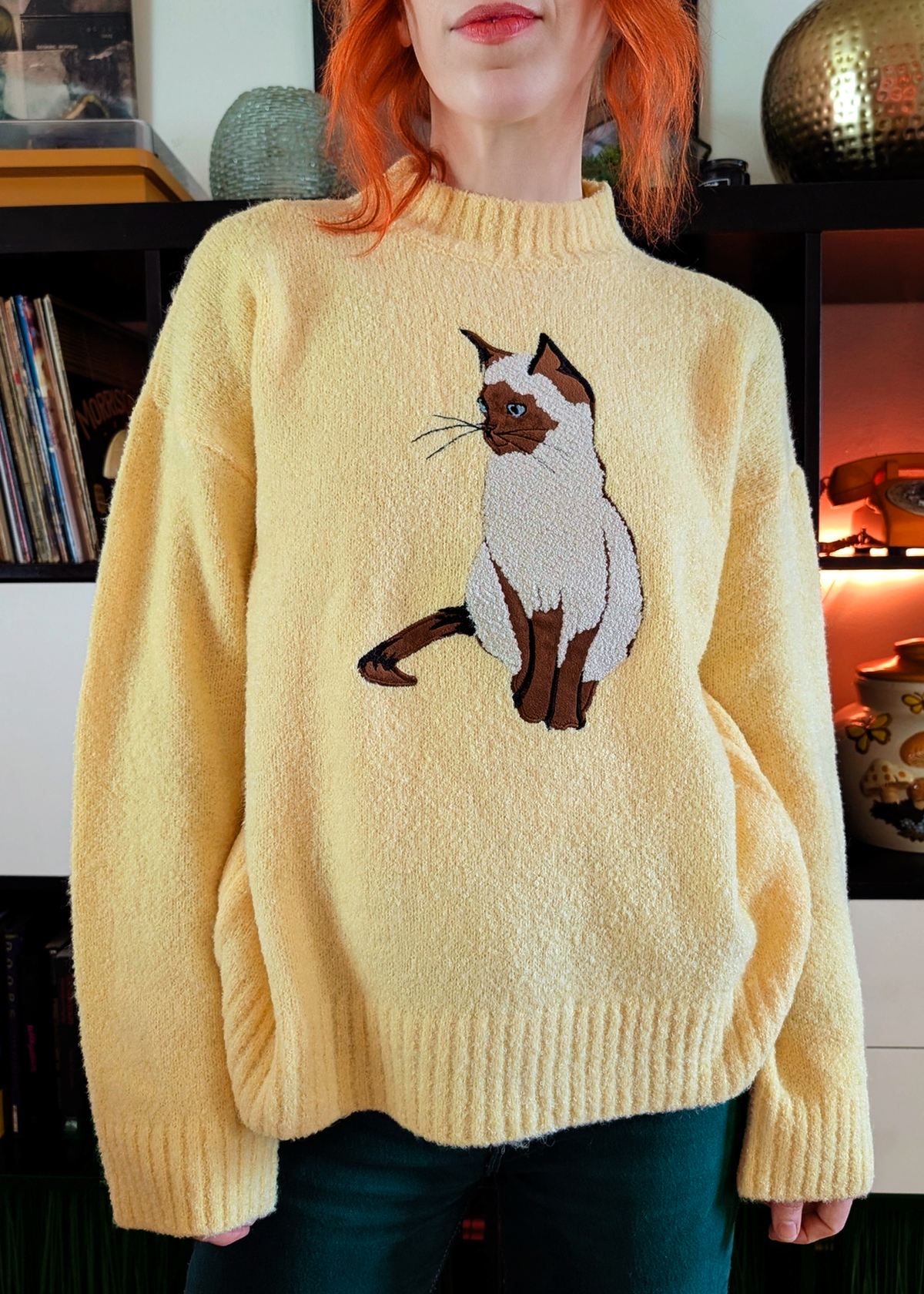 A woman wearing a fuzzy pale yellow knit sweater with a large embroidered cat motif on the front, paired with dark pants, standing indoors in front of record shelves.