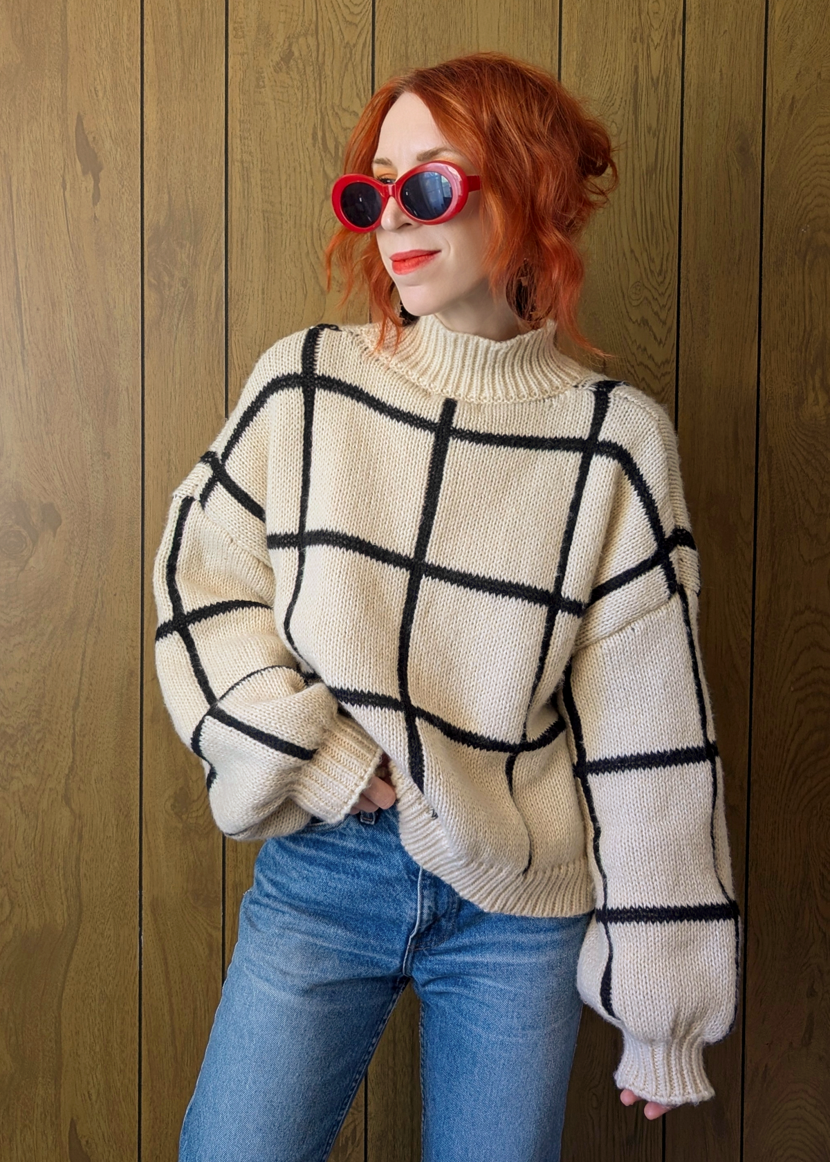 Person wearing a cream sweater with black grid pattern and blue jeans against a wooden background