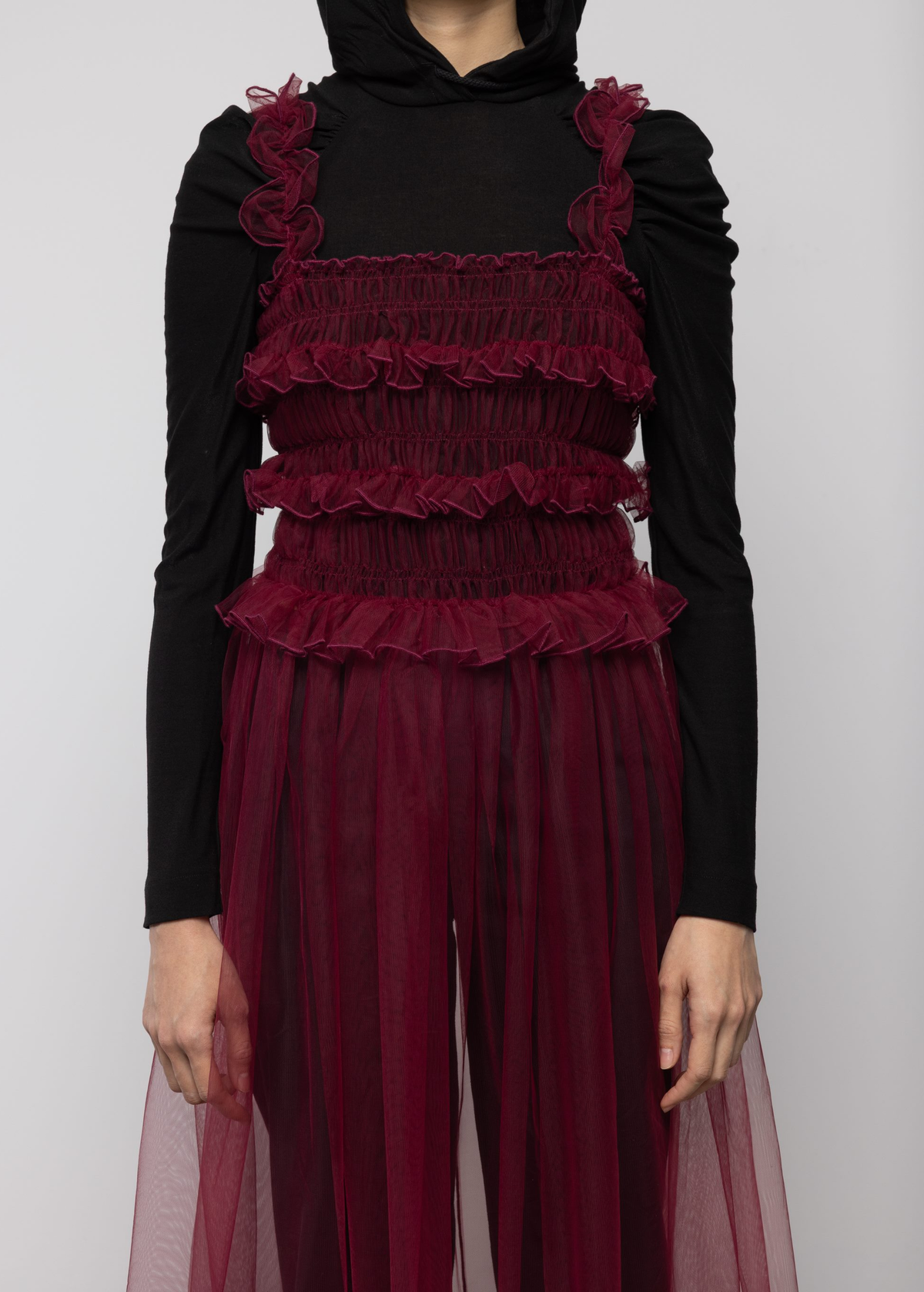 Woman wearing a sheer burgundy tulle dress over a black top and black trousers against a white background