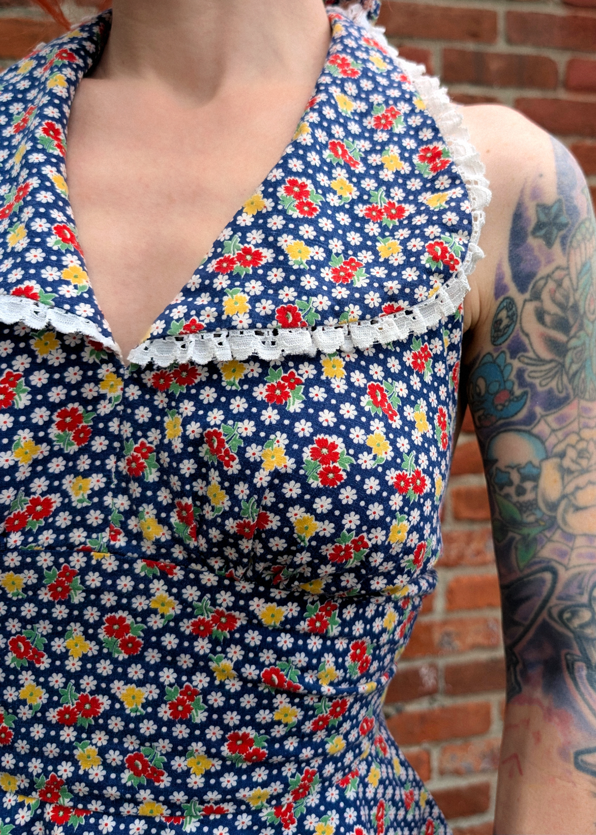 Person wearing a vintage floral halter top with eyelet trim collar standing against a brick wall background