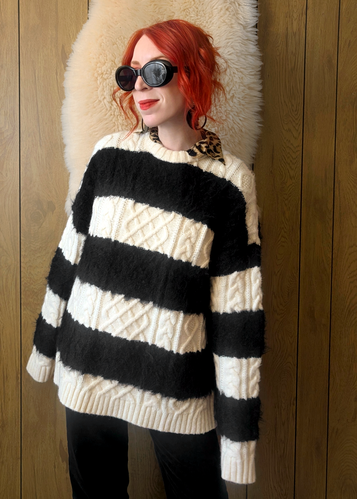 Person wearing a black and cream striped cable knit sweater layered over a leopard print collared shirt, posed against a wooden wall.