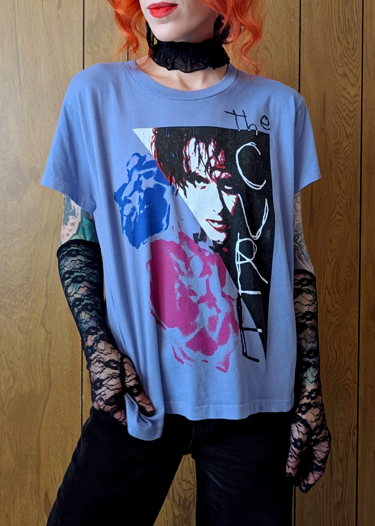 Person wearing a graphic The Cure band t-shirt with a design of Robert Smith, standing against a wooden panel background.