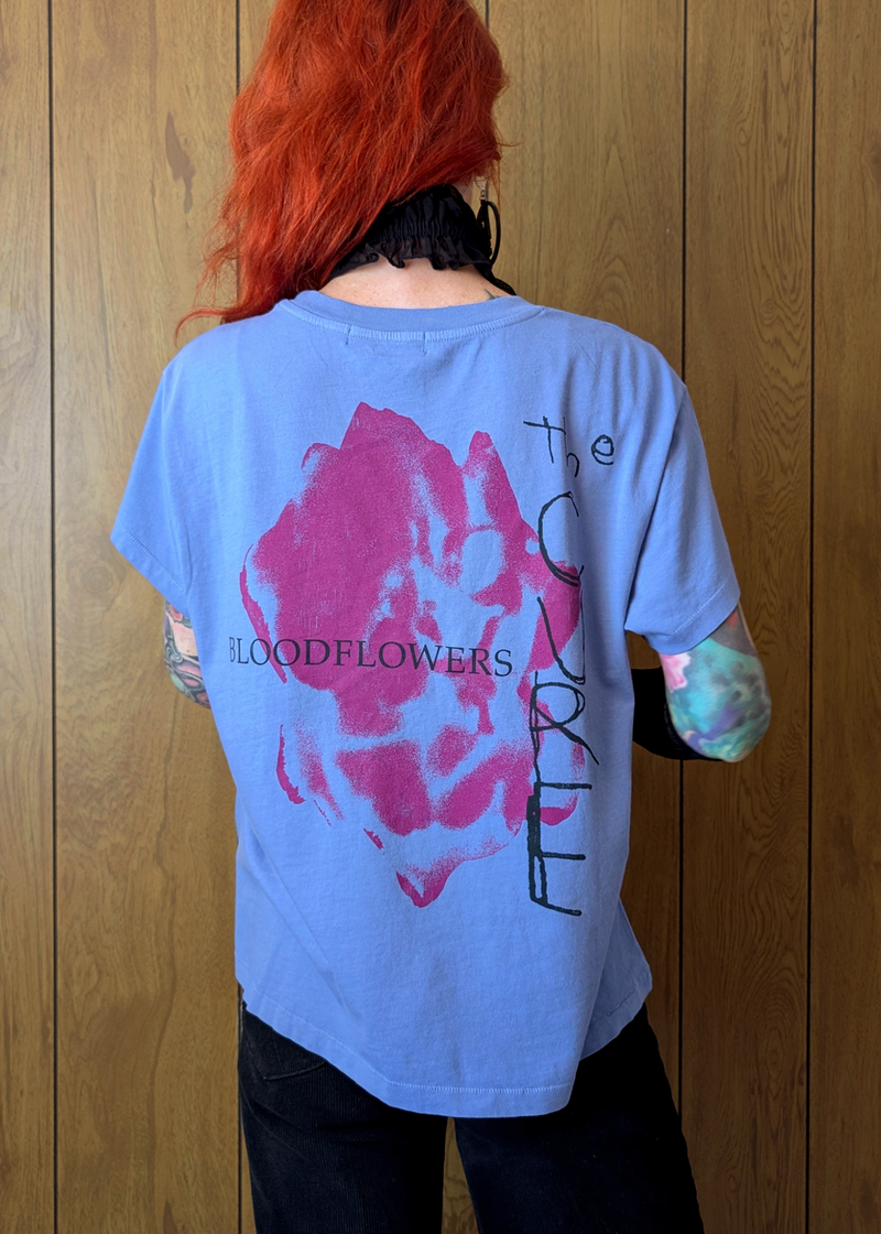 Person wearing a blue t-shirt with a graphic design and text, standing against a wooden panel background.