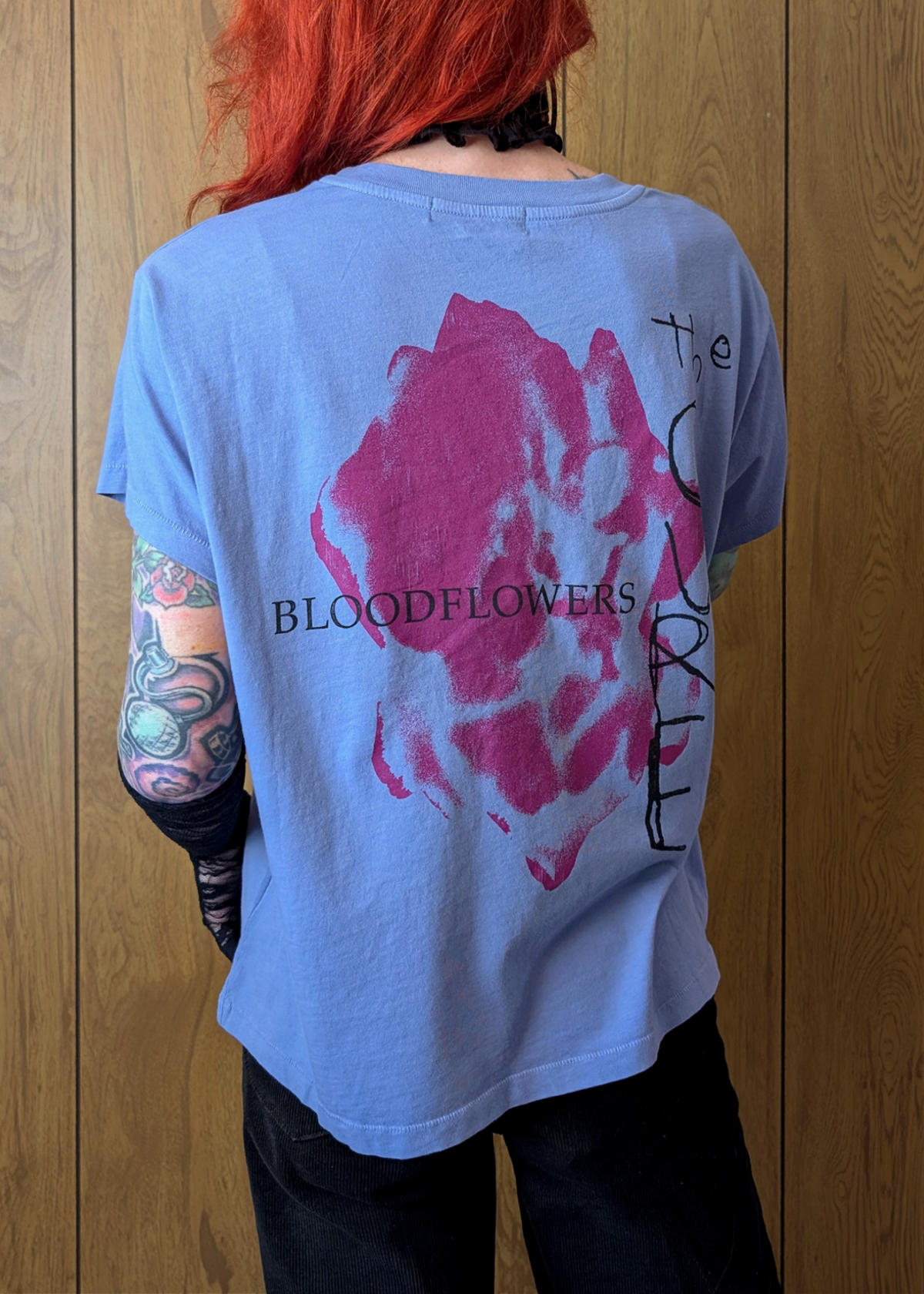 Back view of a person wearing a periwinkle blue  The Cure t-shirt with a pink graphic and Bloodflowers text, standing against a wooden wall.