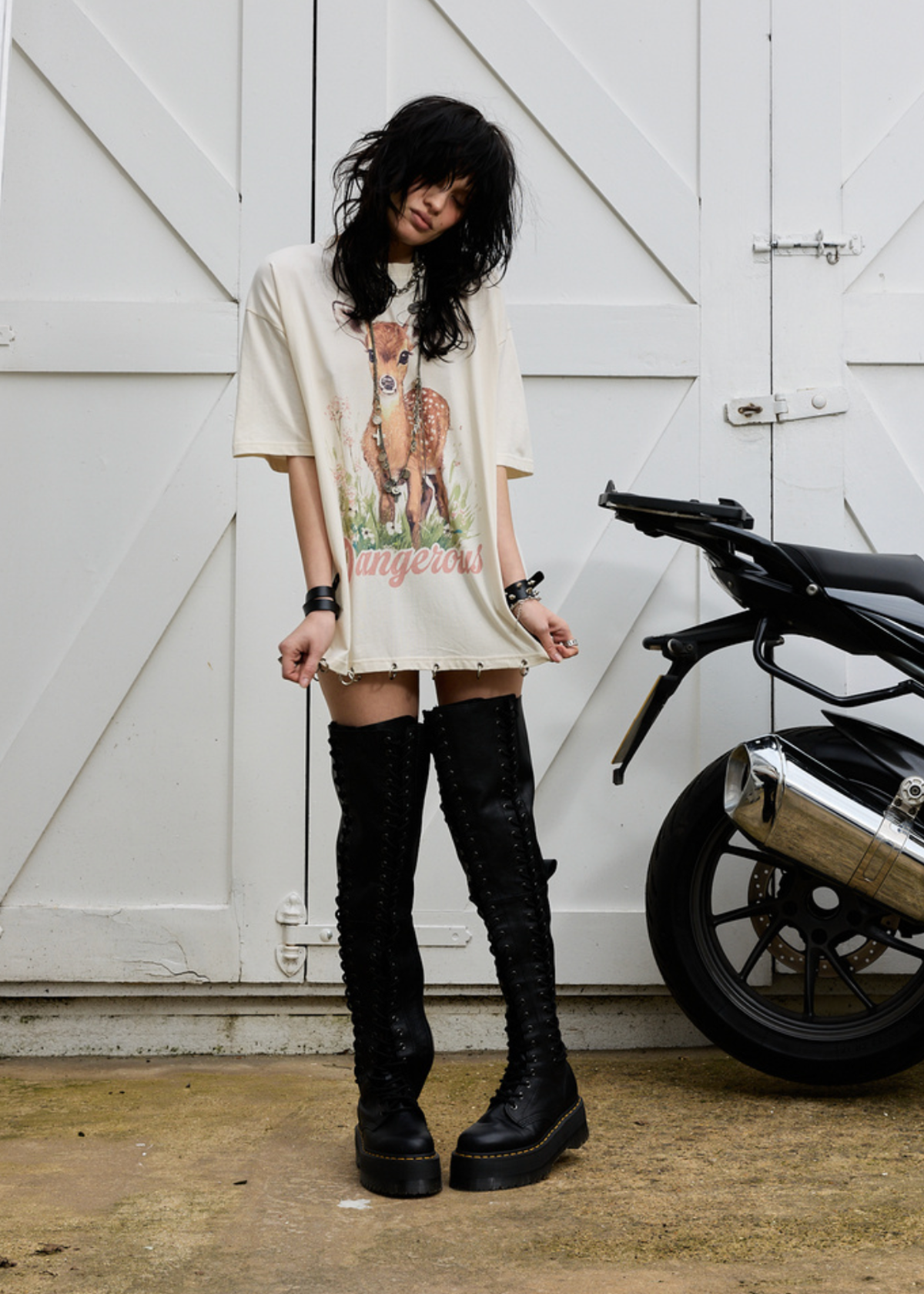 Person wearing a Deer graphic t-shirt and knee-high boots standing next to a motorcycle against a white wooden door.