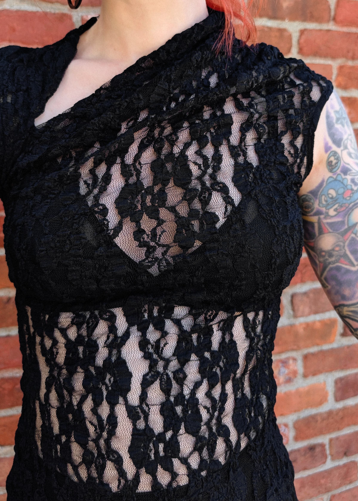A close-up of a person wearing a black lace top with sheer floral lace panels and a draped neckline, standing against a red brick wall.