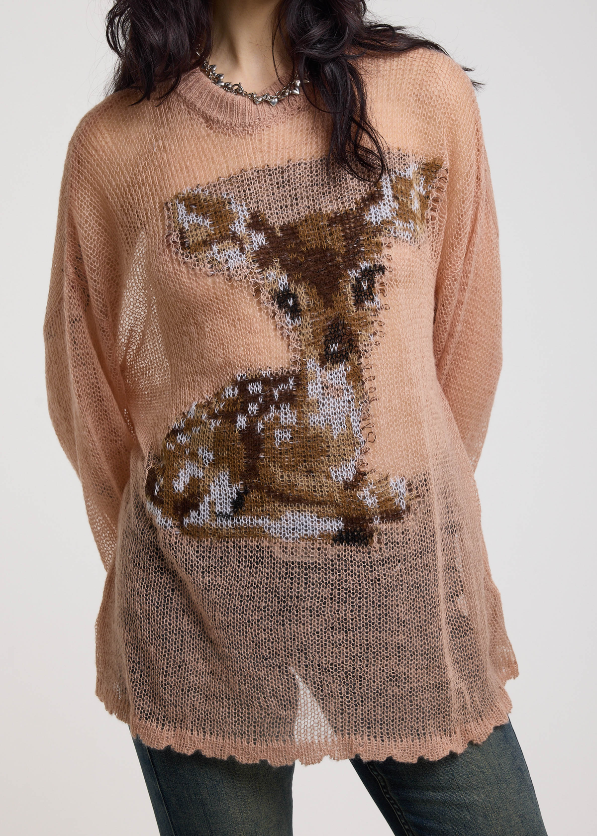 Woman wearing a dusty pink open knit lightweight sweater with a deer design on a plain background