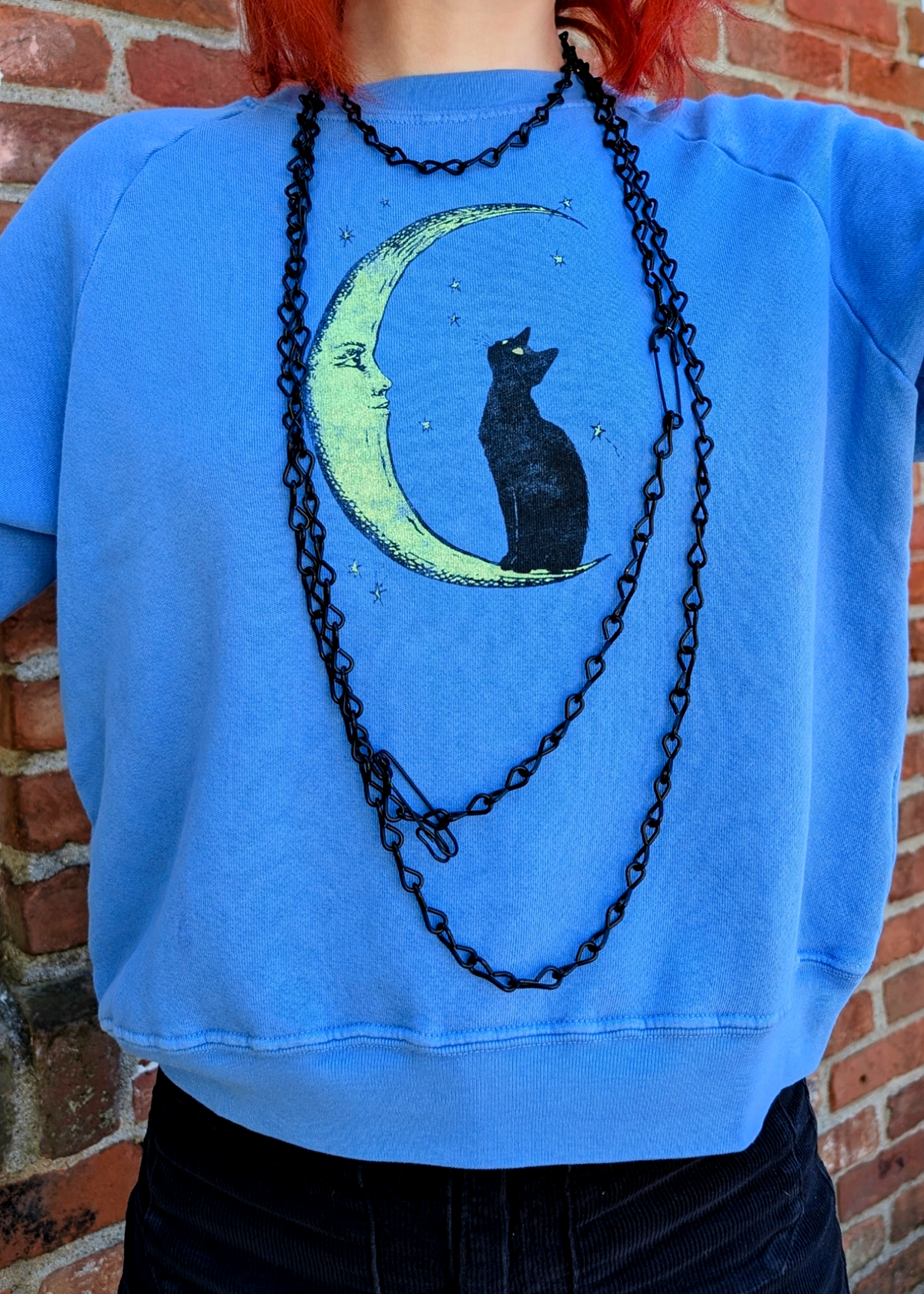 A person wearing a bright blue cropped sweatshirt with a graphic of a black cat sitting on a crescent moon, layered with a long black chain necklace, standing against a red brick wall.