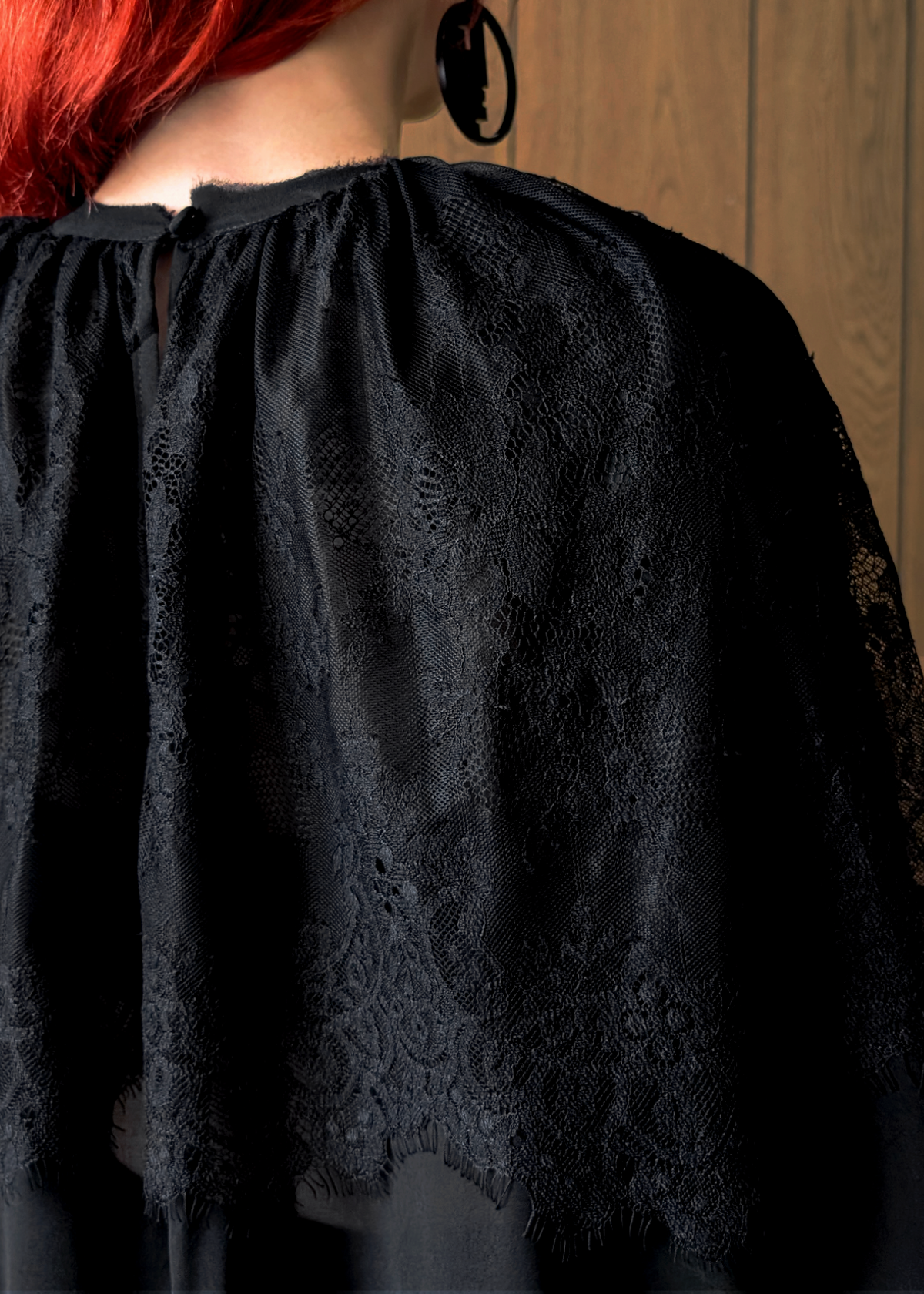 Black lace top worn by a person with red hair, against a wooden panel background