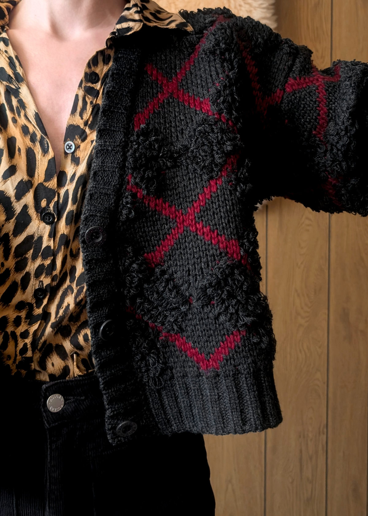 details of a charcoal and red argyle "x" patterned cardigan over a leopard print shirt against a wooden wall.