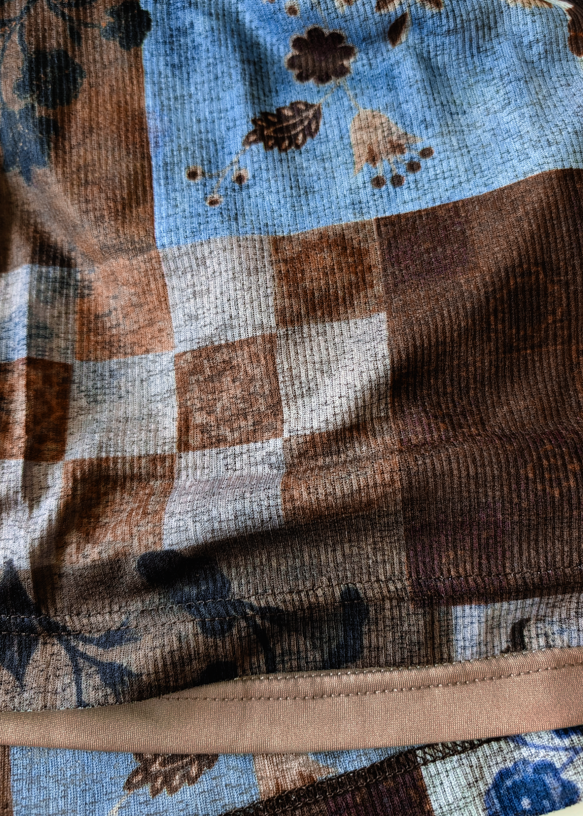 A close-up of a patchwork-patterned fabric with a mix of brown, beige, and light blue textured sections, including a checkered pattern, floral motifs, and a solid beige trim.