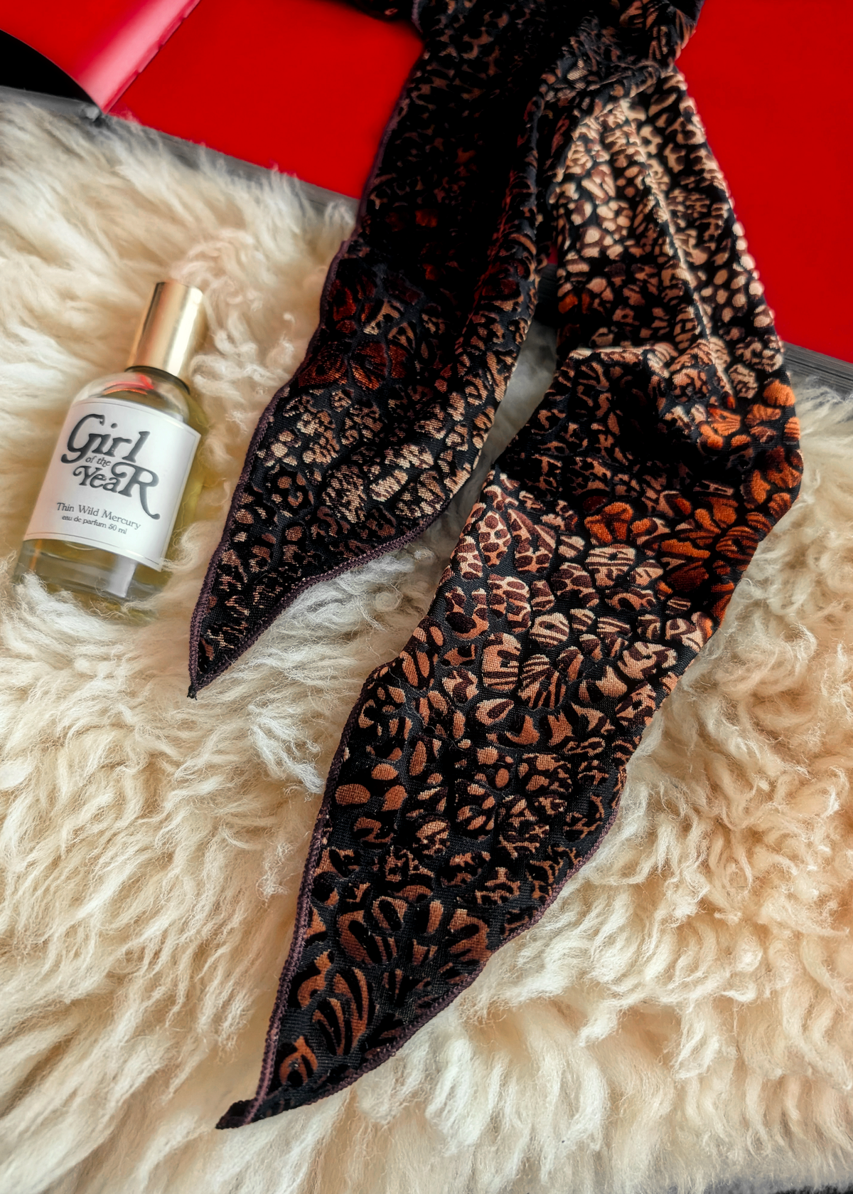 Lifestyle photo of a animal print burnout velvet scarf laid flat on a cream faux fur rug with a red book and bottle of perfume