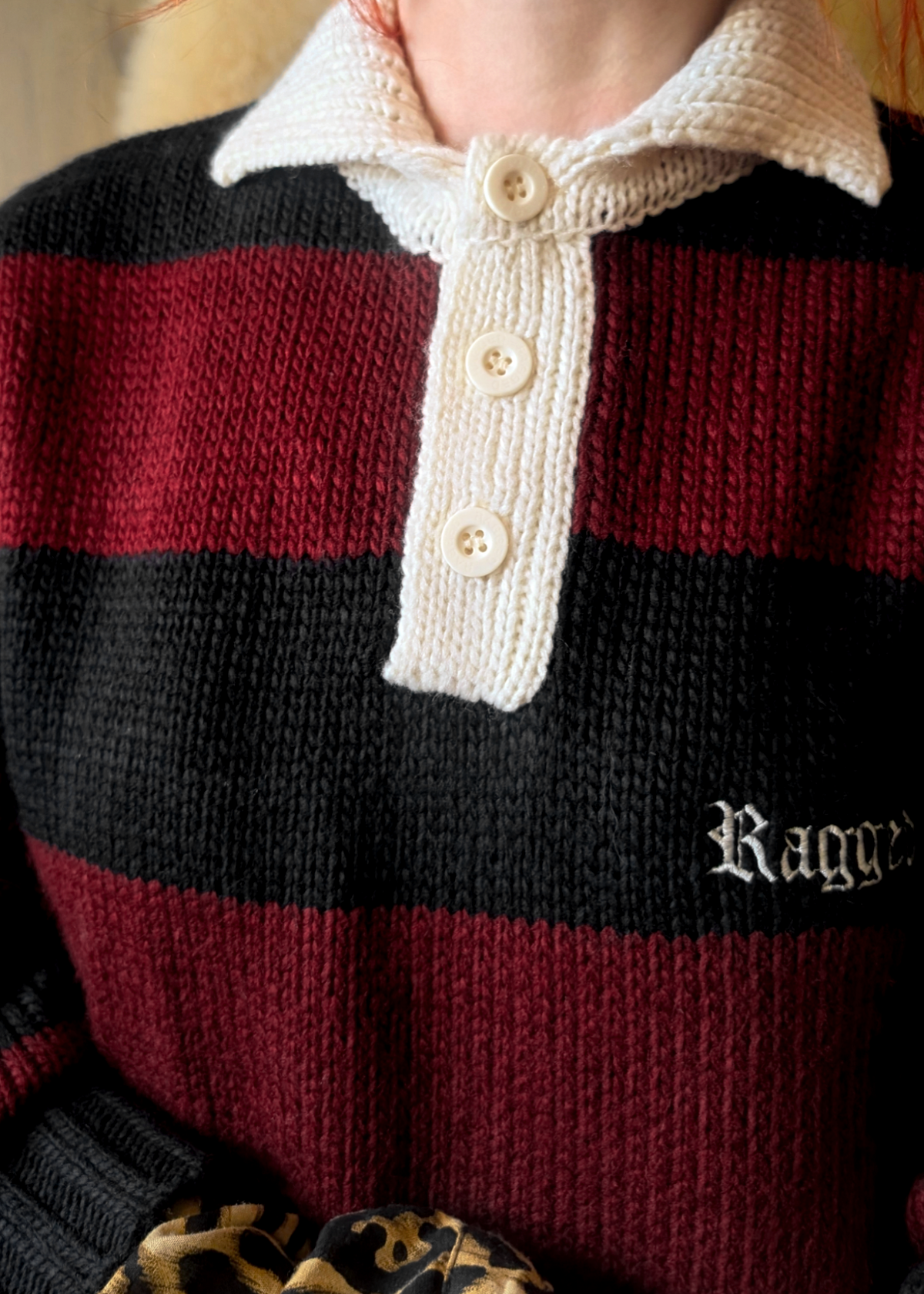Close-up of a person wearing a red and black striped polo sweater with a cream collar and buttons, featuring the brand 'Ragged' embroidered at chest