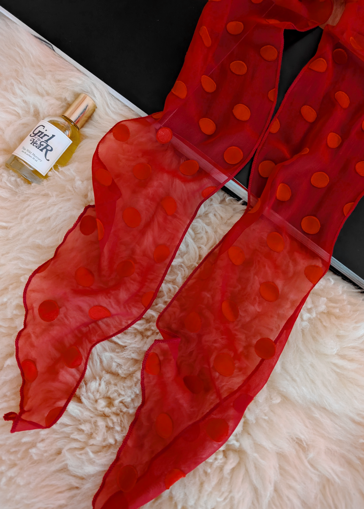 Lifestyle photo of a red polka dot scarf laid flat on a cream faux fur rug with a black book and bottle of perfume