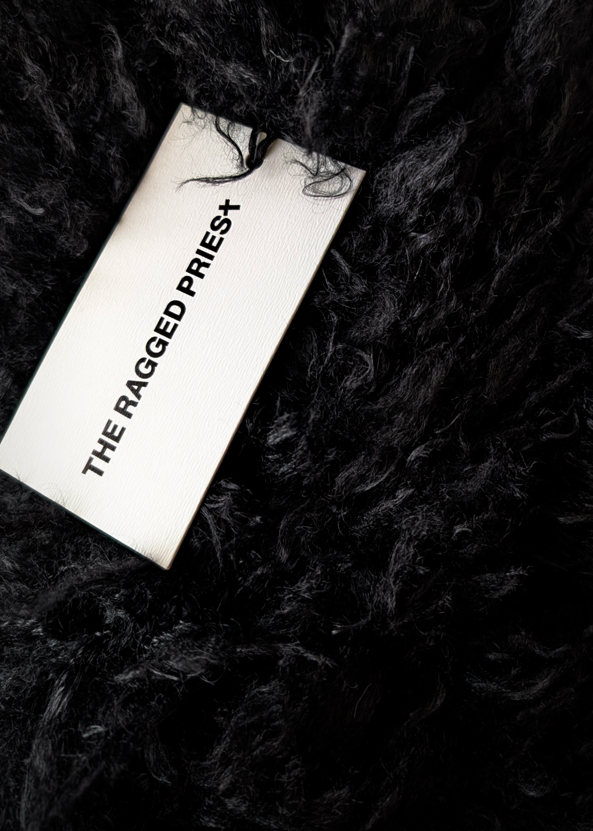 A close-up of a black shaggy curly faux-fur scarf with a white tag reading 'THE RAGGED PRIEST