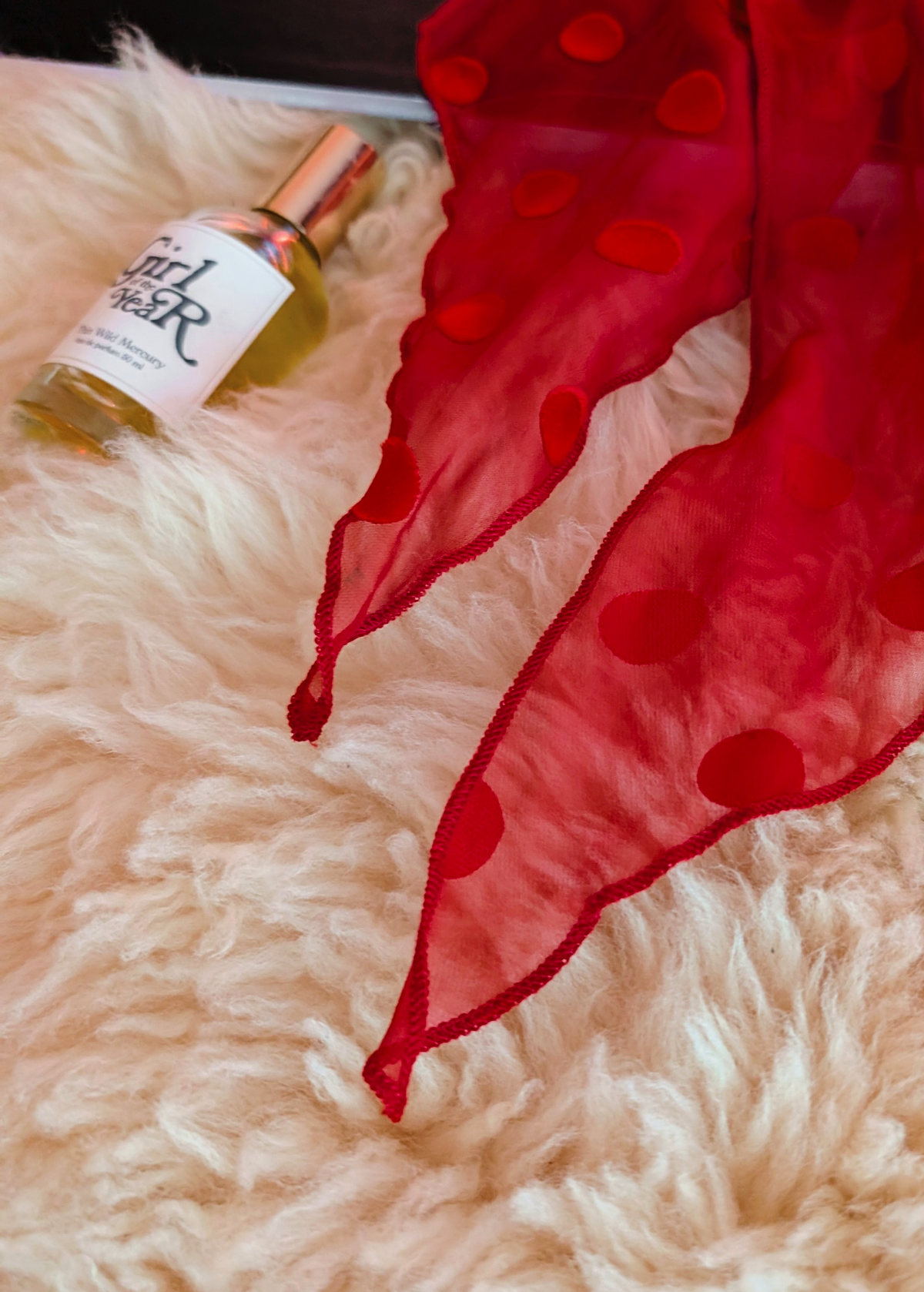 Lifestyle photo of a red polka dot scarf laid flat on a cream faux fur rug with a black book and bottle of perfume