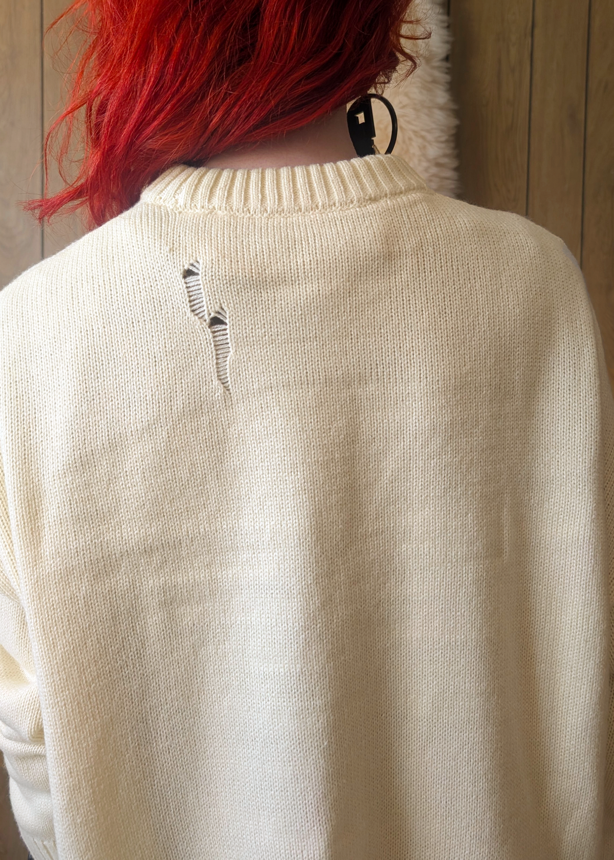Person wearing a cream sweater with an intentional destroyed detail on the back, standing against a wooden wall.