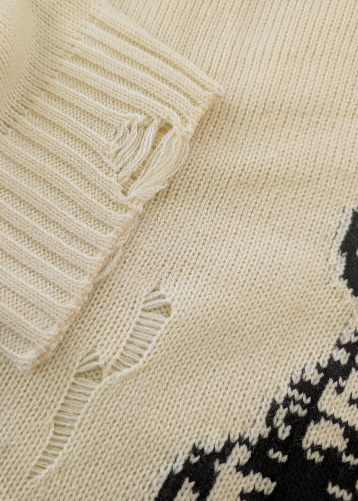 Close-up of a beige knitted sweater with black pattern and details of the intentional distressed details on the sleeves and front of sweater 