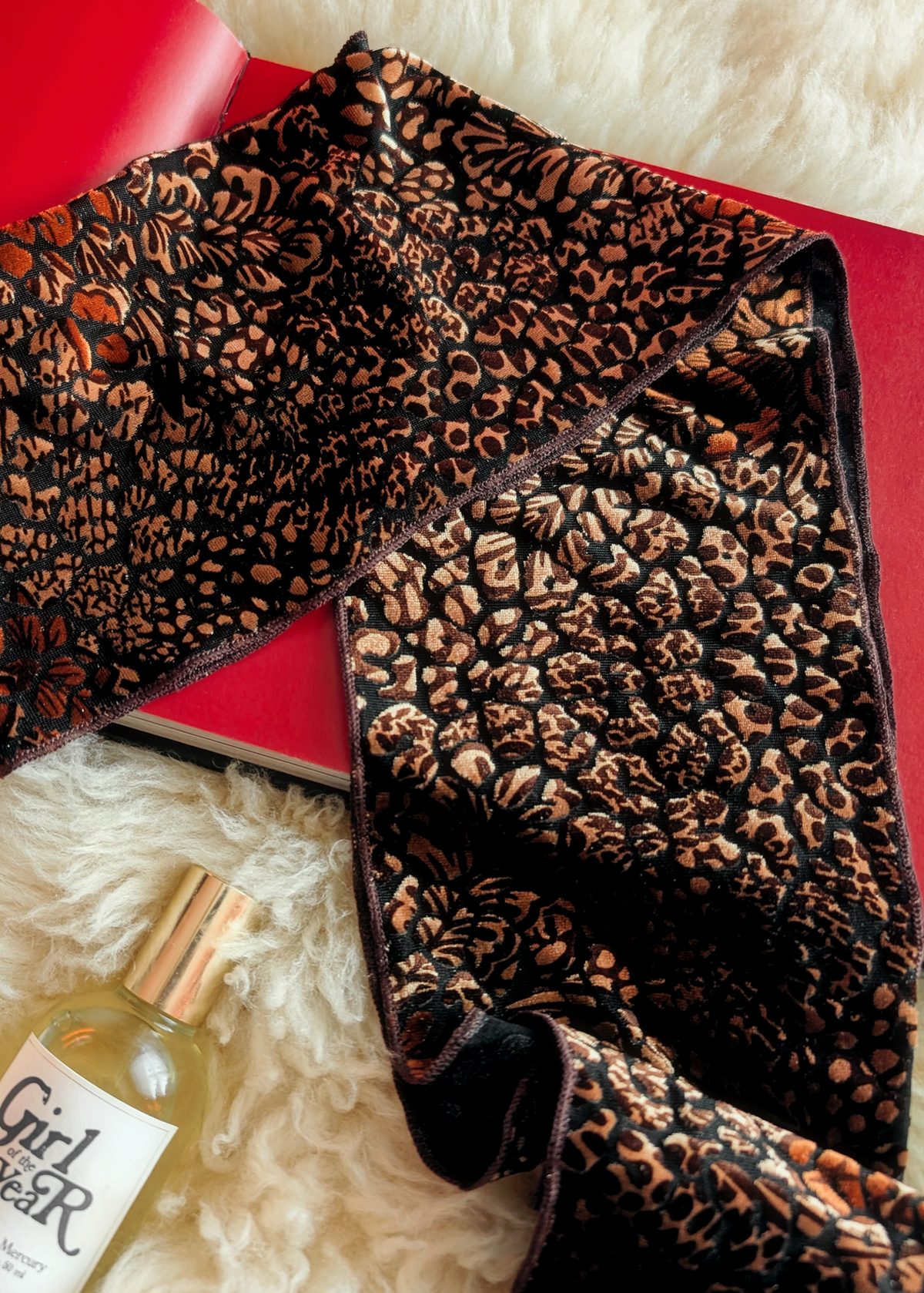 Lifestyle photo of a animal print burnout velvet scarf laid flat on a cream faux fur rug with a red book and bottle of perfume