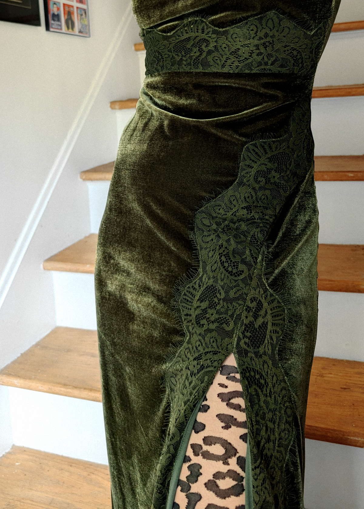 An olive green velvet dress with a lace panel and a high slit showing leopard print tights, photographed in an indoor setting with wooden stairs in the background.