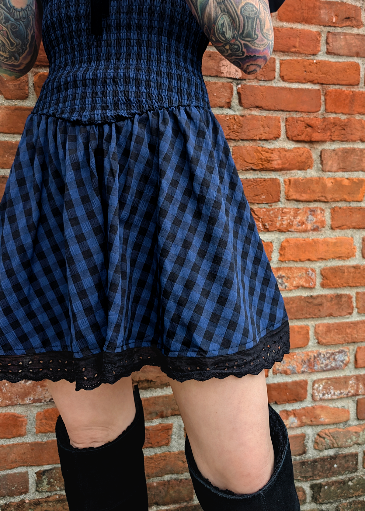 Person wearing a blue and black checkered dress with lace trim against a brick wall.