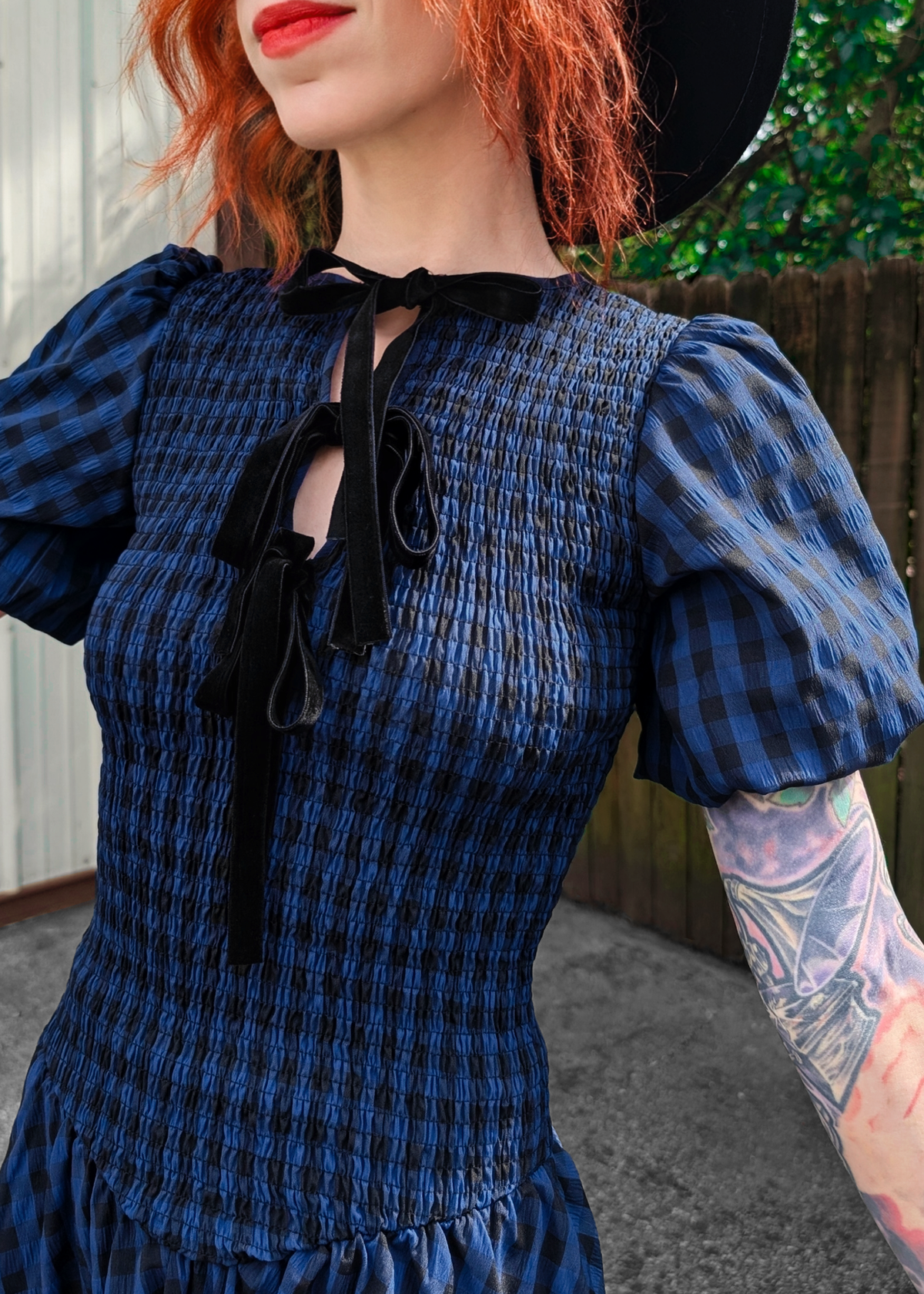 Person wearing a blue and black checkered dress with puffed sleeves and a black hat.