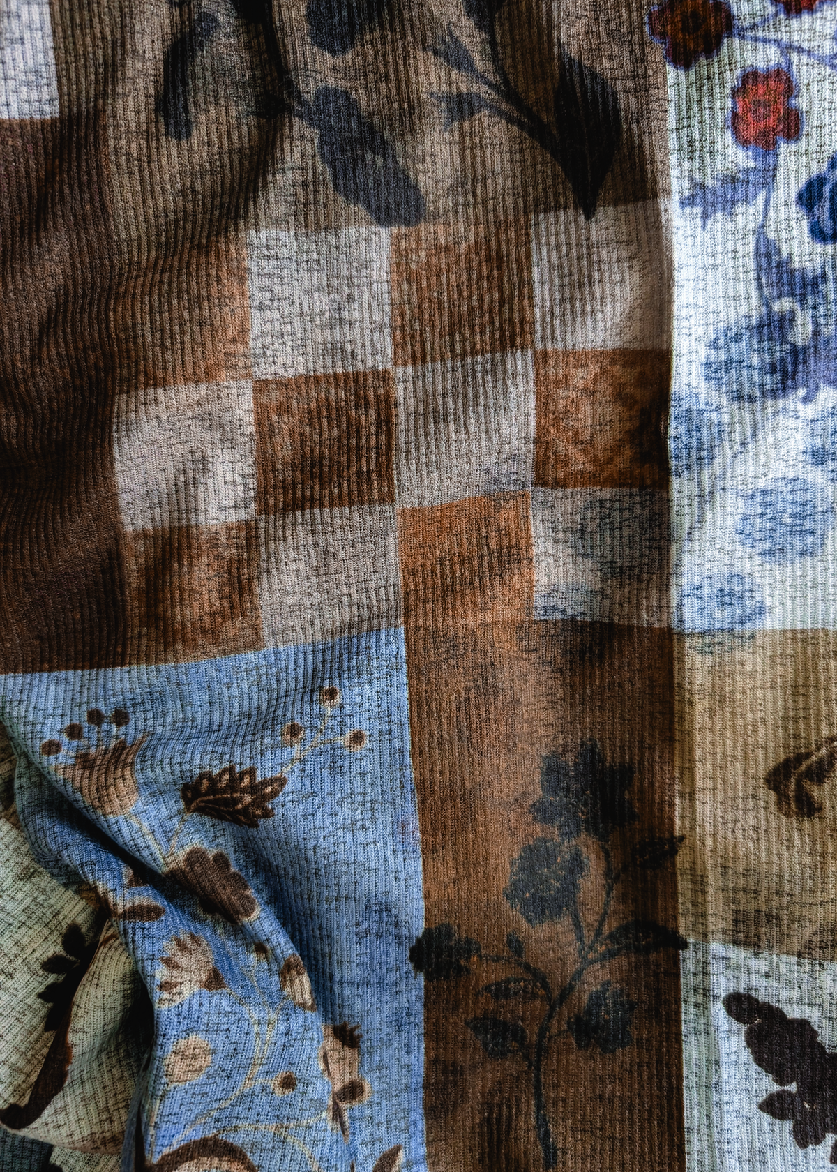 A close-up of a patchwork printed fabric with a rib texture, featuring blocks of brown, beige, and light blue with floral and botanical patterns.