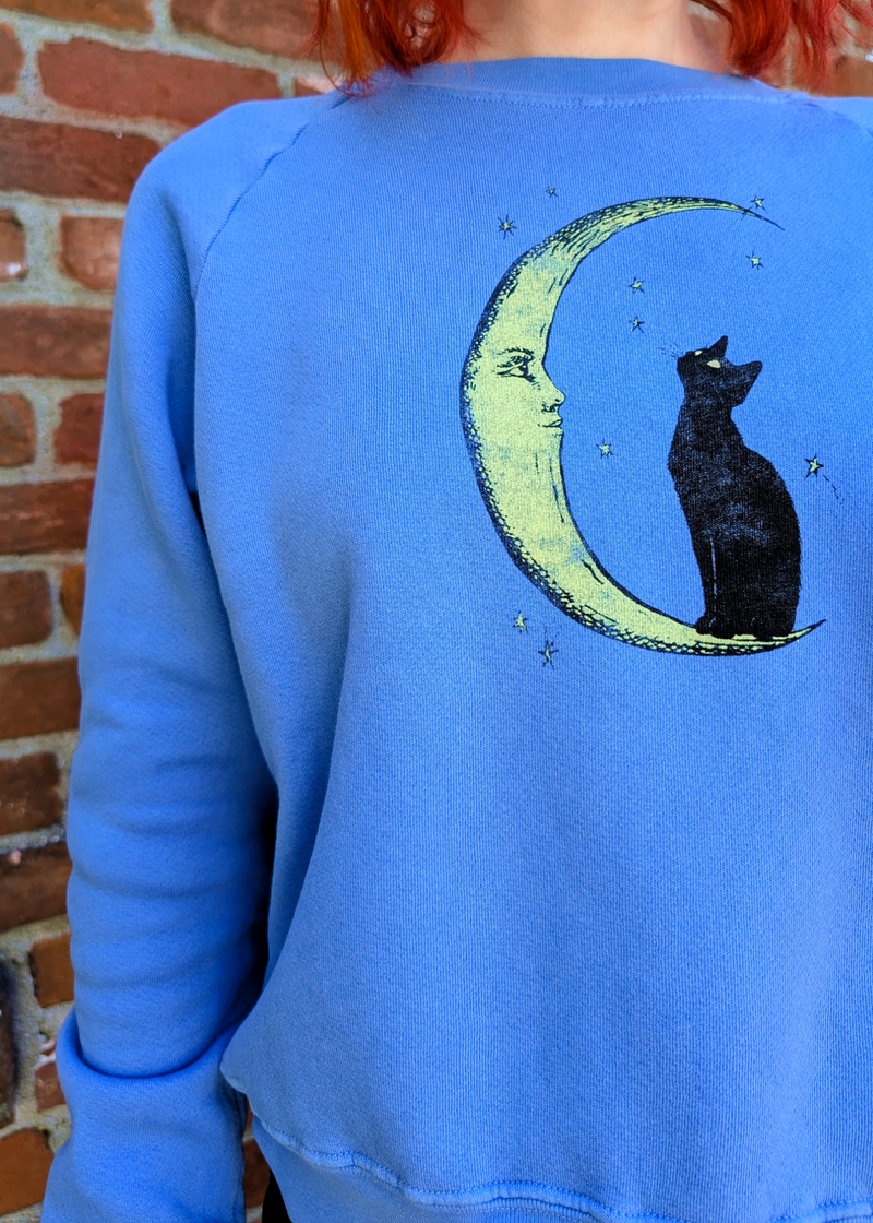 A person wearing a bright blue sweatshirt with a graphic of a black cat sitting on a crescent moon, photographed outdoors against a brick wall.