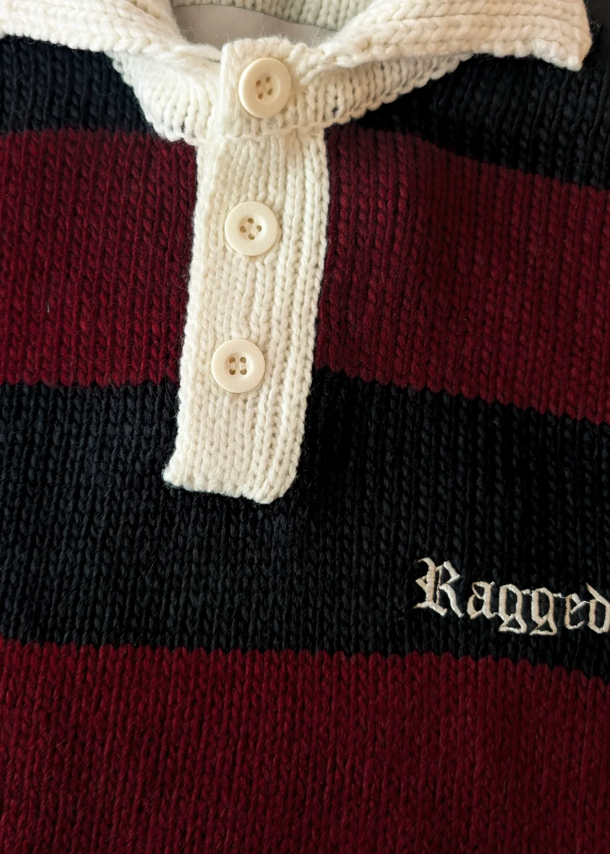 Close-up of a striped sweater with a buttoned collar and 'Ragged' branding.