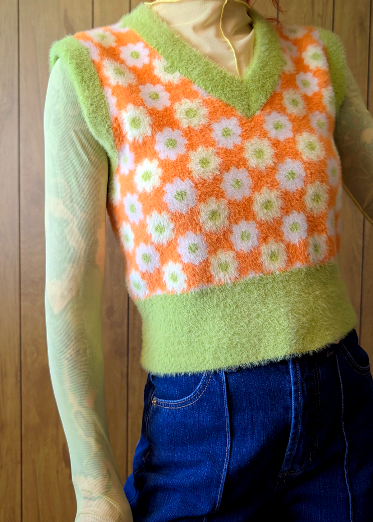 person wearing a colorful knitted vest with a pattern of orange, white, and green flowers against a wooden background.