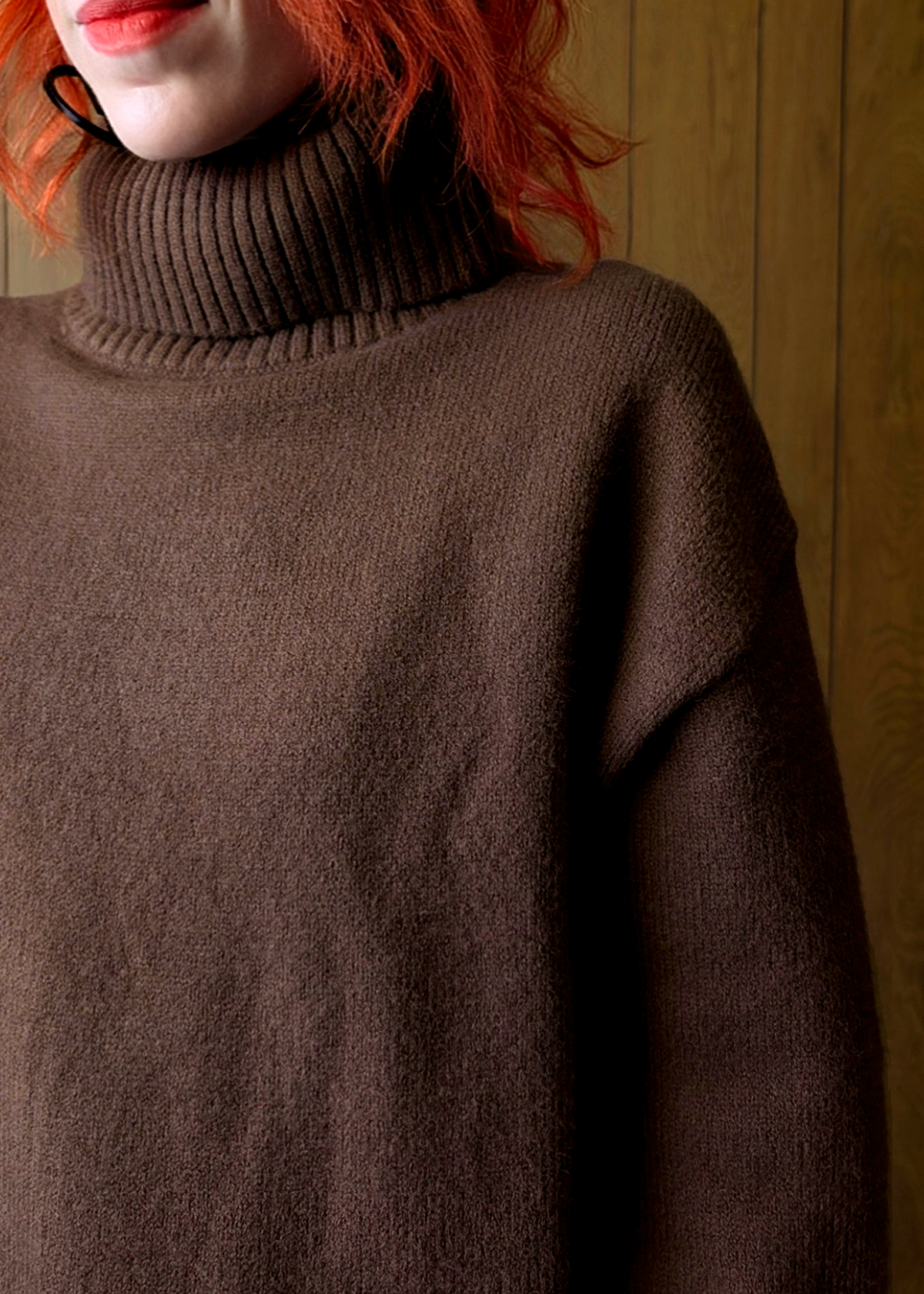 Brown sweater worn by a person with red hair against a wooden panel background