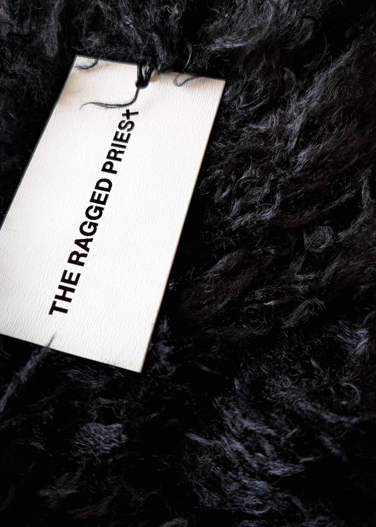 A  close-up of a black shaggy curly faux-fur scarf with a white tag reading 'THE RAGGED PRIEST