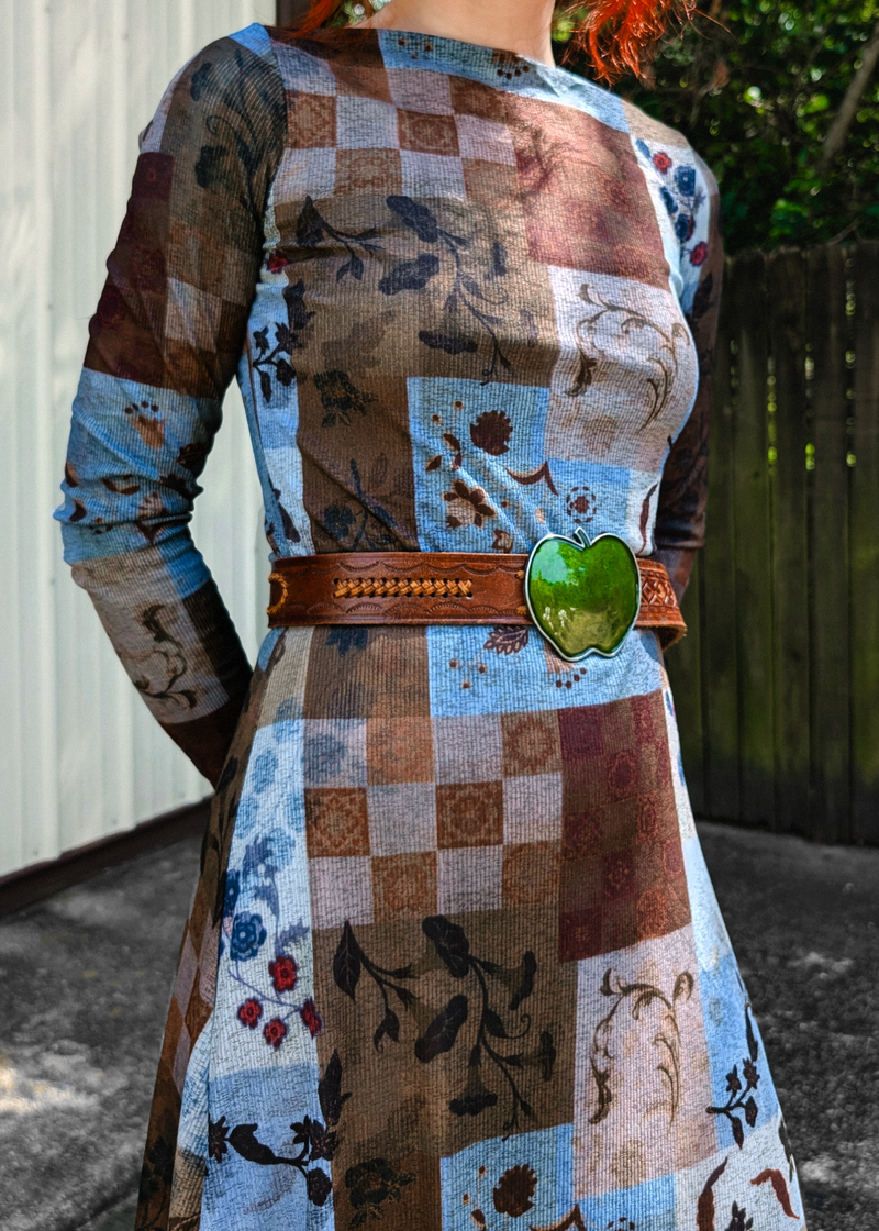 A woman wearing a long-sleeve patchwork print dress with brown, beige, and light blue floral and checkerboard patterns, styled with a brown leather belt and a green apple-shaped buckle, photographed outdoors.