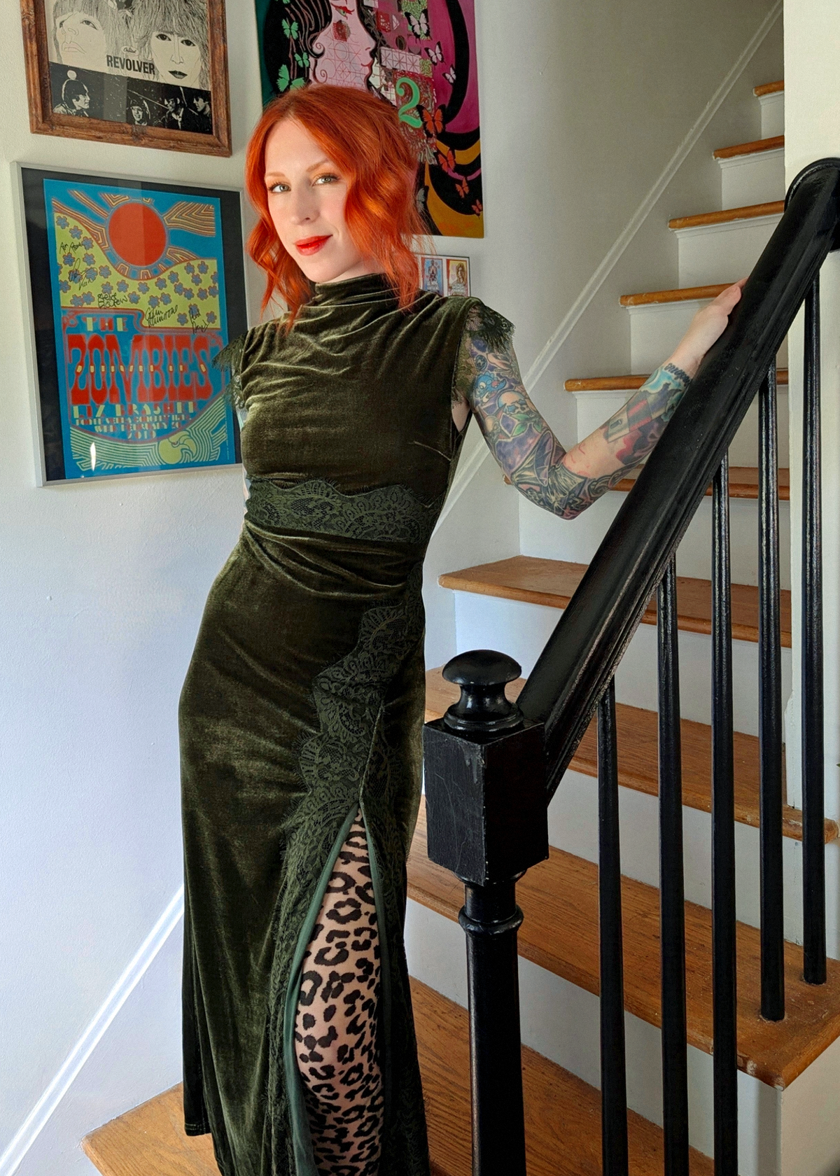 A woman with red hair stands on wooden stairs holding a black handrail, wearing a dark olive green velvet sleeveless dress with lace detailing, styled with leopard print tights and burgundy velvet heels. Framed posters hang on the wall behind her.