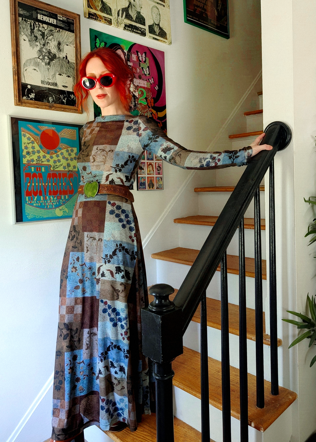 A woman with red hair and red sunglasses stands on wooden stairs holding a black handrail, wearing a long-sleeved patchwork print maxi dress with blue, brown, and beige tones.
