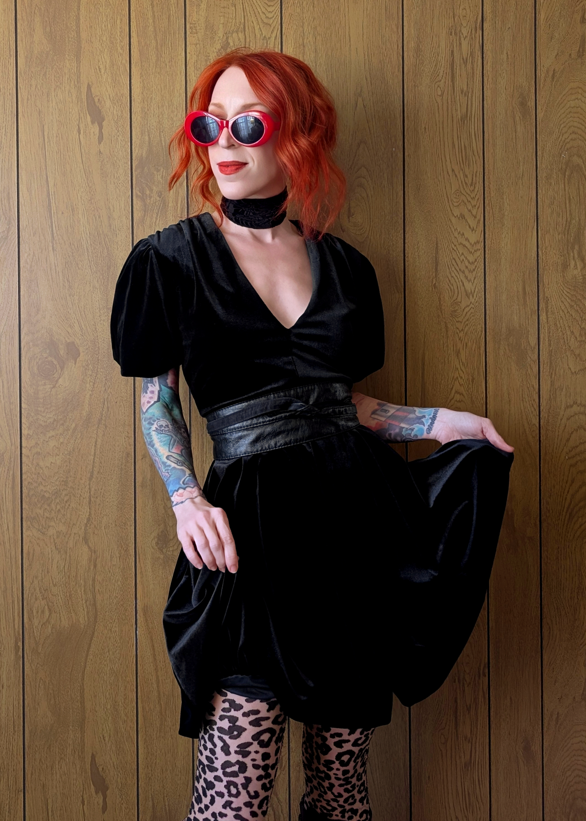 Person with red hair wearing a black velvet mini dress and leopard print tights against a wooden wall.