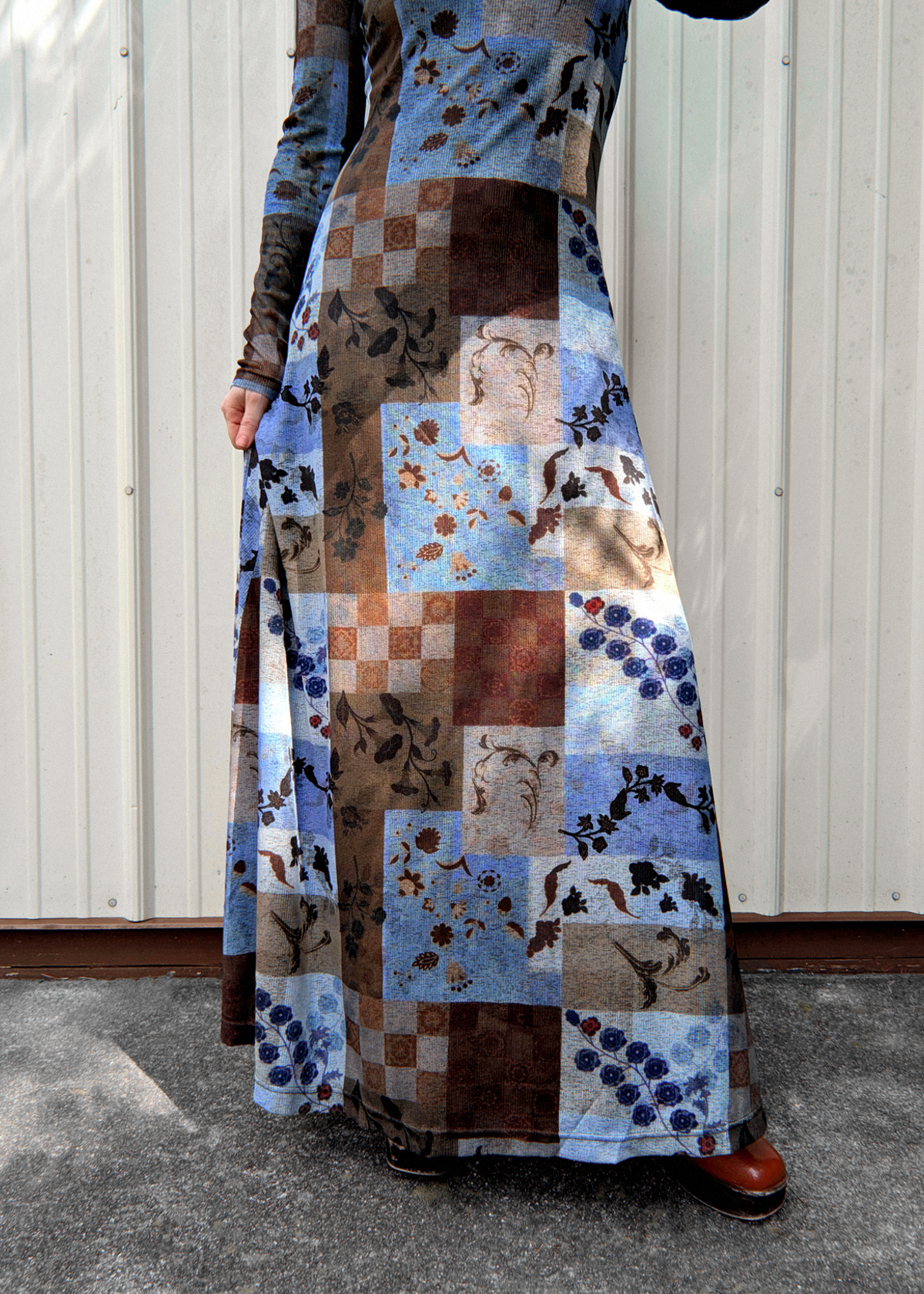 A person wearing a long-sleeved maxi dress with a rustic patchwork print in shades of blue, brown, and beige, standing against a white corrugated metal wall.