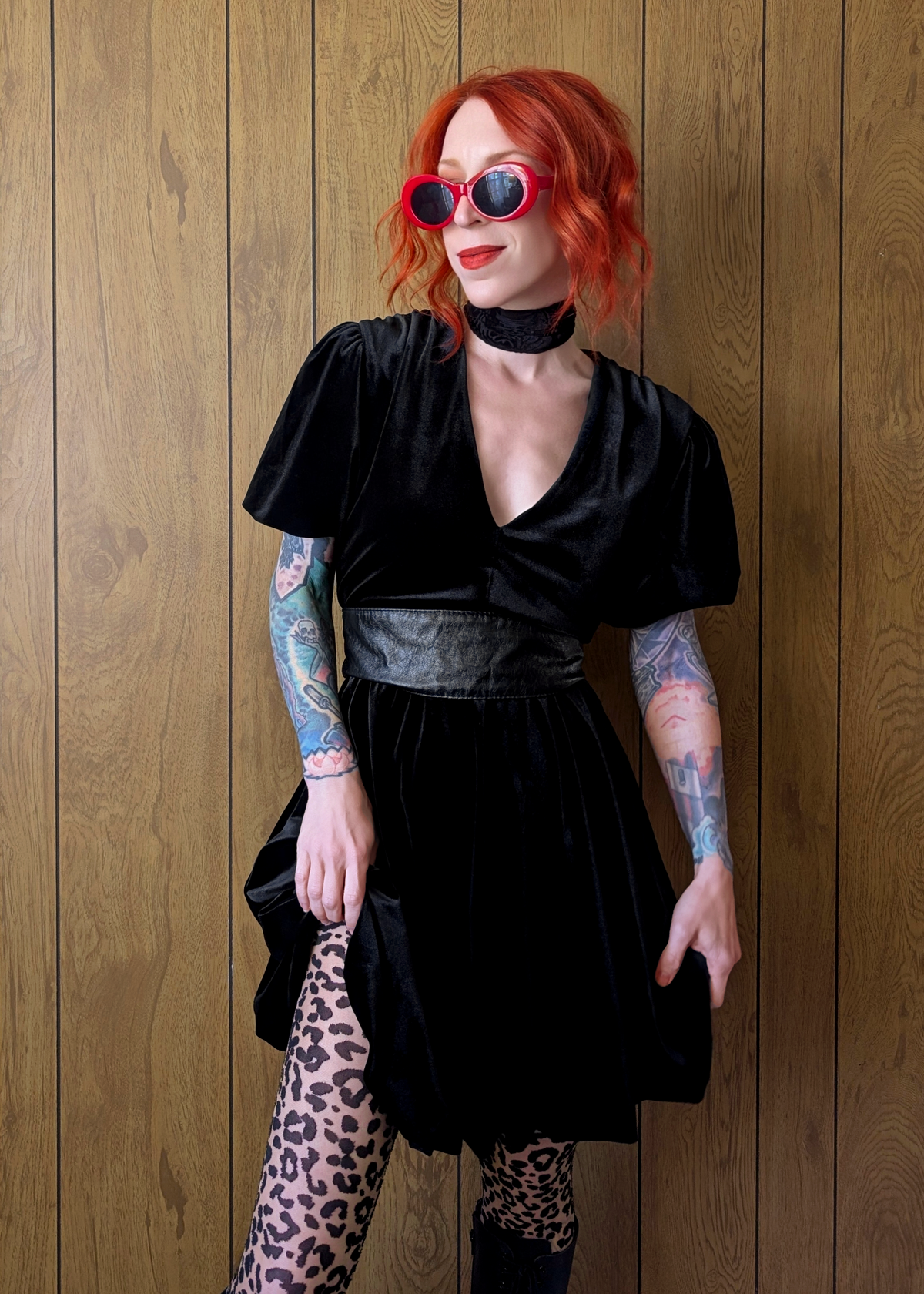 Person wearing a black velvet mini dress with leopard print tights against a wooden wall.