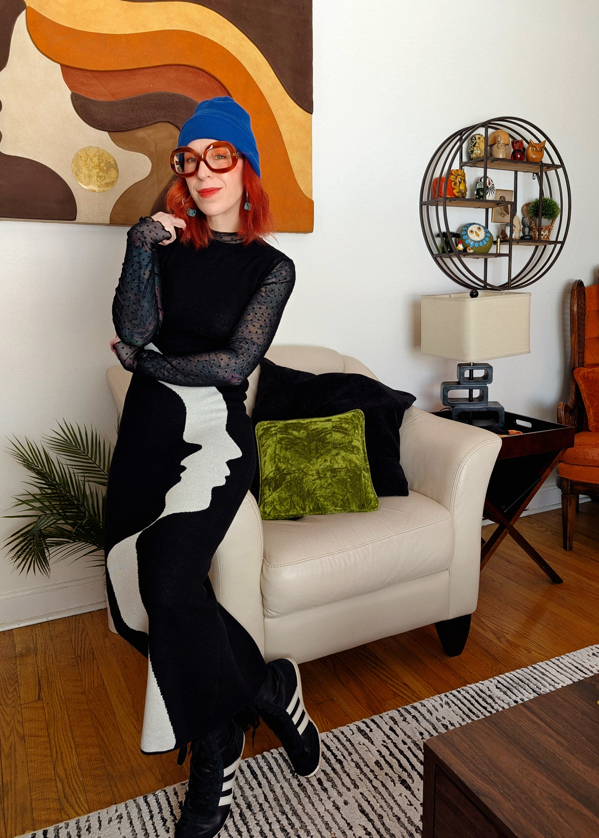 Person sitting on a white chair in a living room with colorful abstract art and decor, wearing a black and white knit dress with face design.